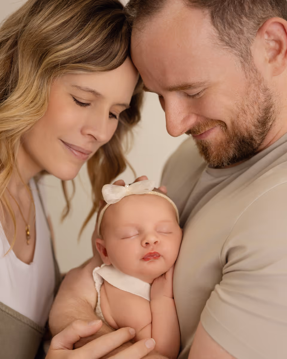 A smiling couple tenderly holding a sleeping baby with a headband and bow.