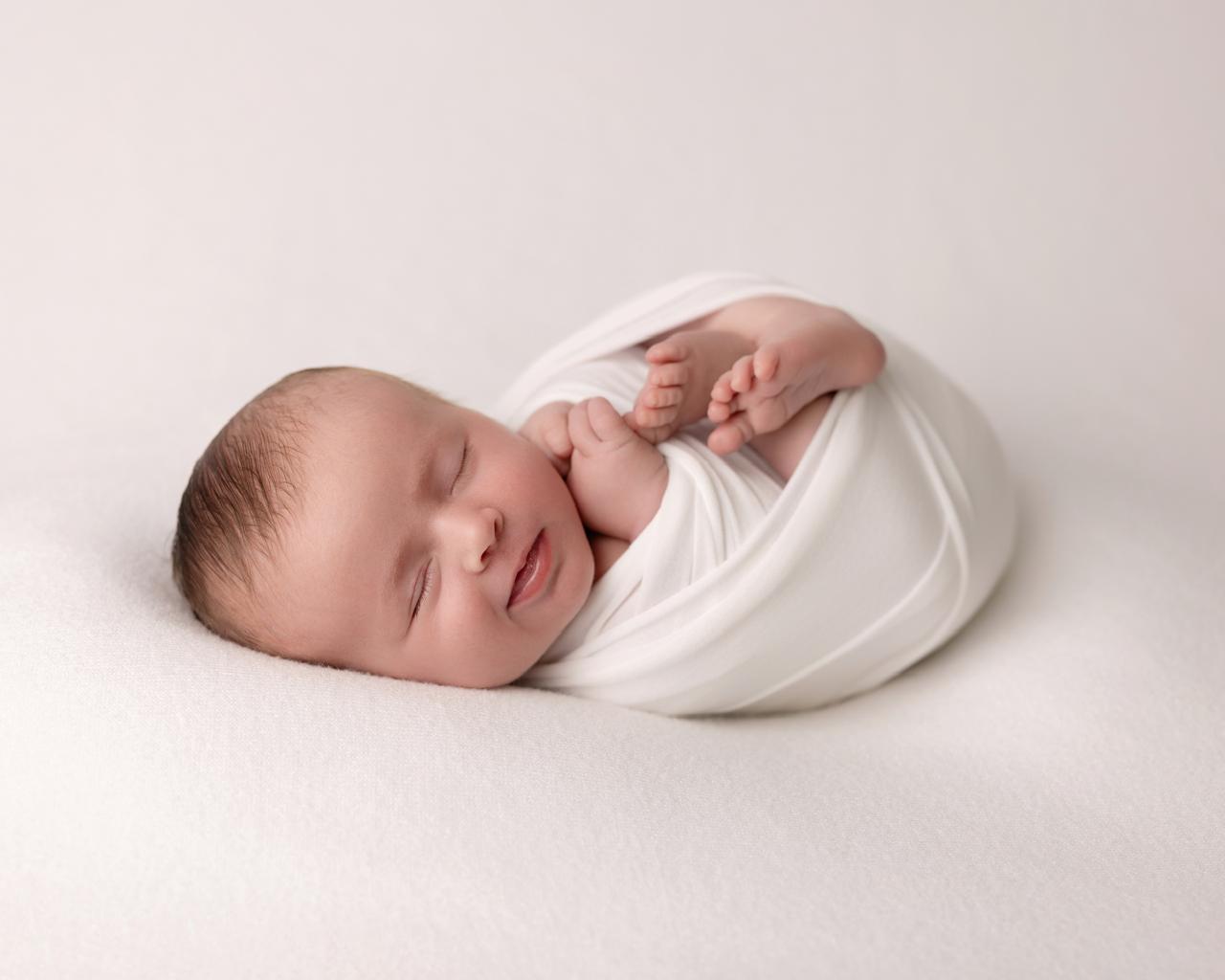Newborn baby peacefully sleeping wrapped in a white cloth on a soft white surface.