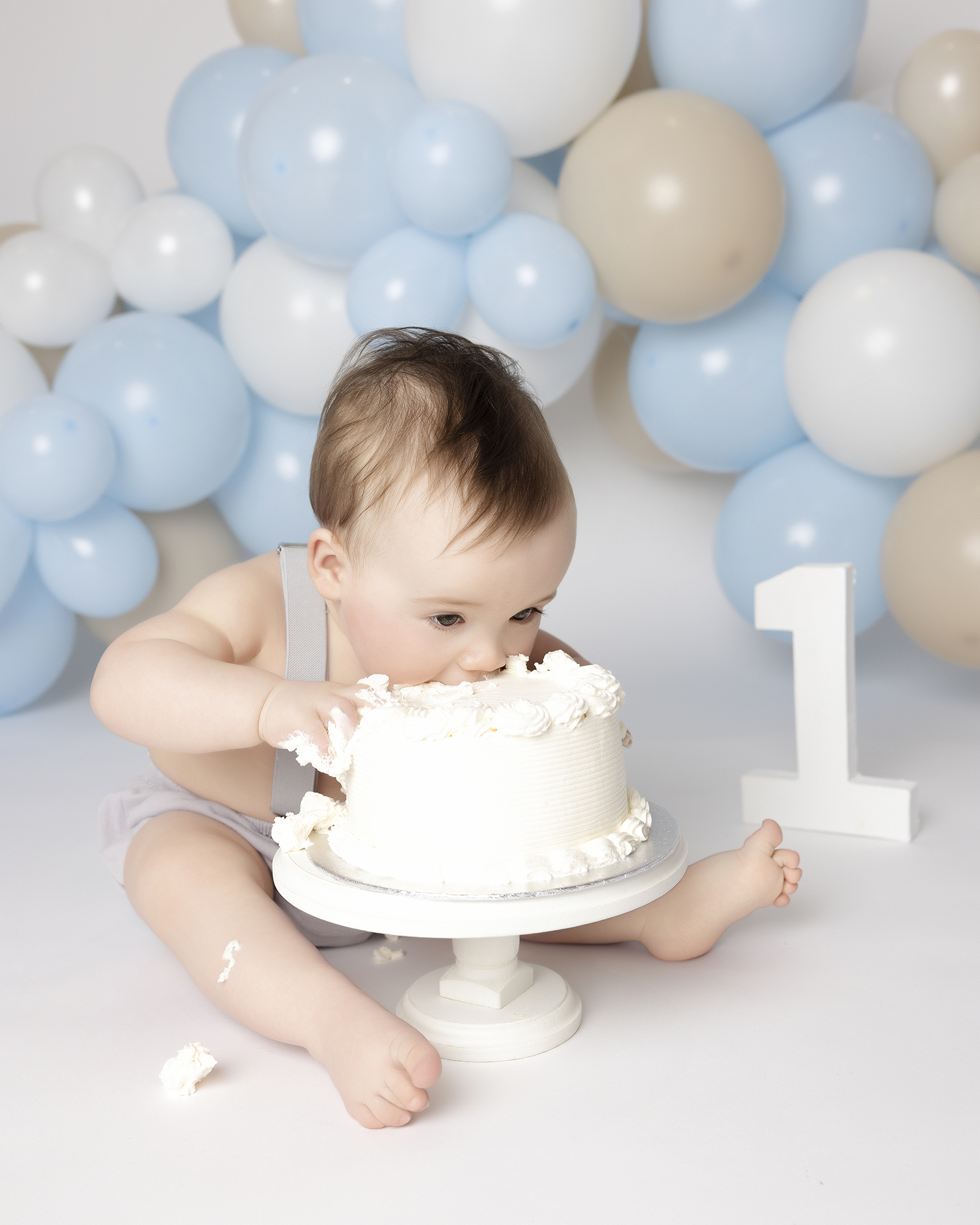 Baby in gray suspenders sitting and eating white frosted cake with blue and beige balloons and a white number 1 decoration in the background.