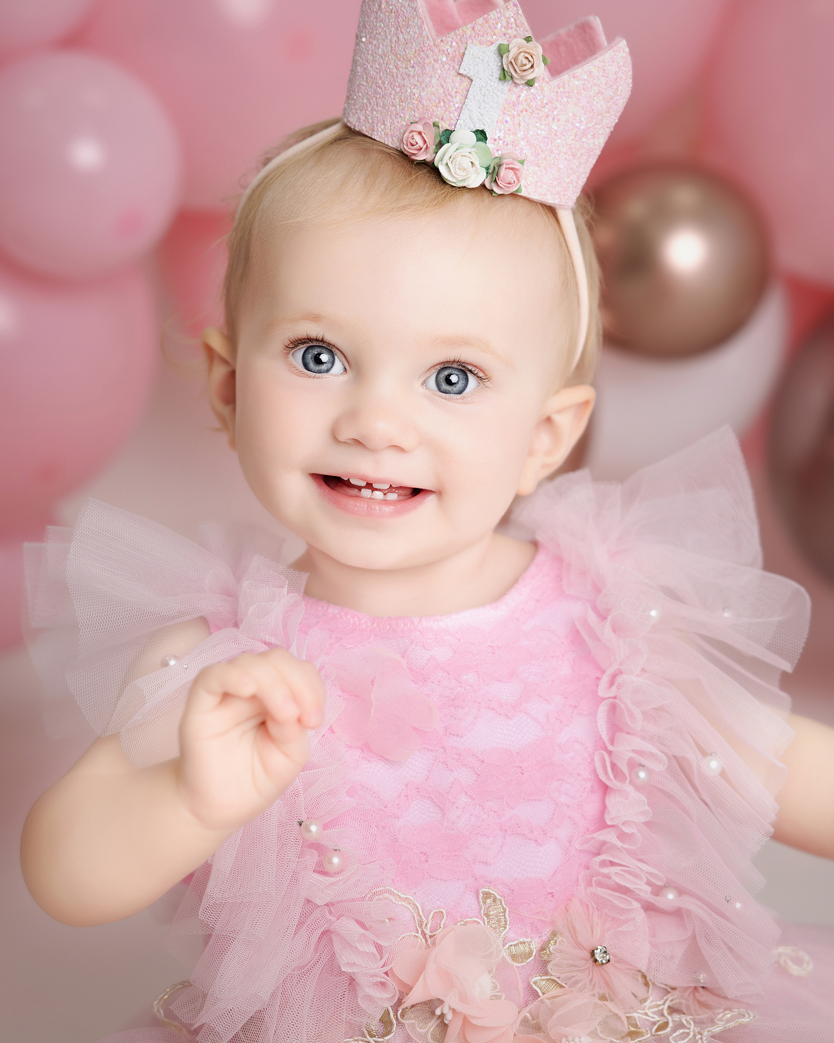Smiling baby girl with blue eyes wearing a pink dress and a glittery pink crown with a number 1 and flower decorations.