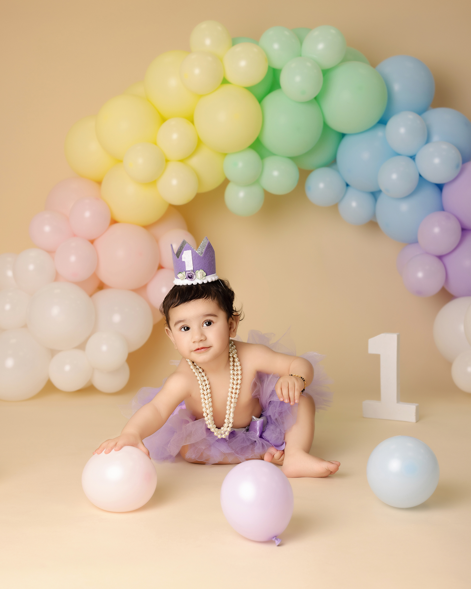 Toddler in a purple tutu and pearl necklace with a purple crown marked '1', sitting on the floor with pastel balloons and a large number one decoration behind.