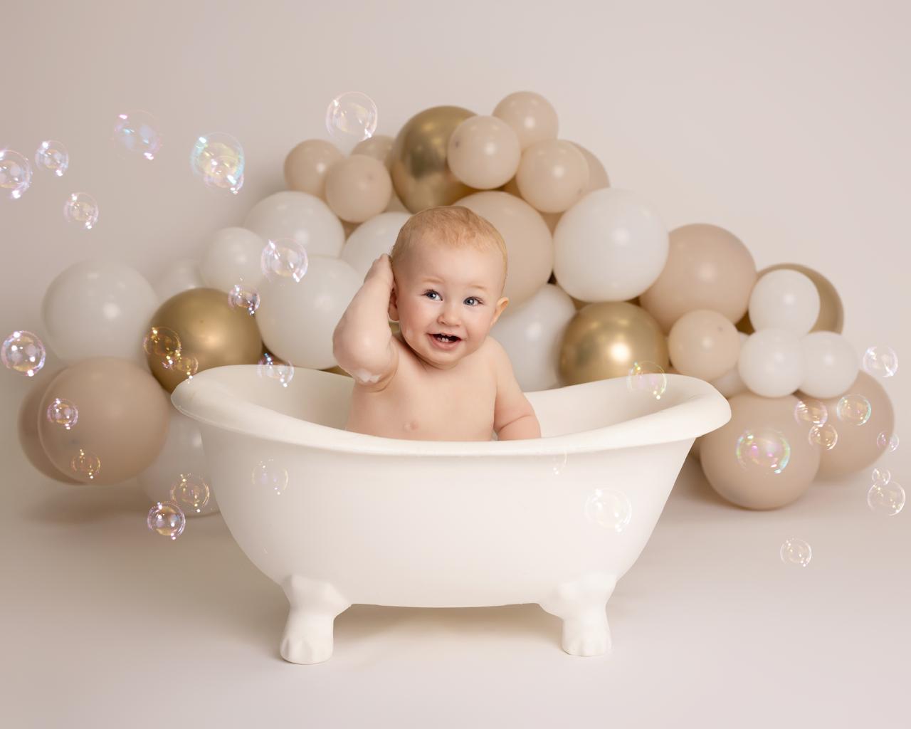 Smiling baby sitting in a white clawfoot bathtub with beige and gold balloons and floating bubbles around.