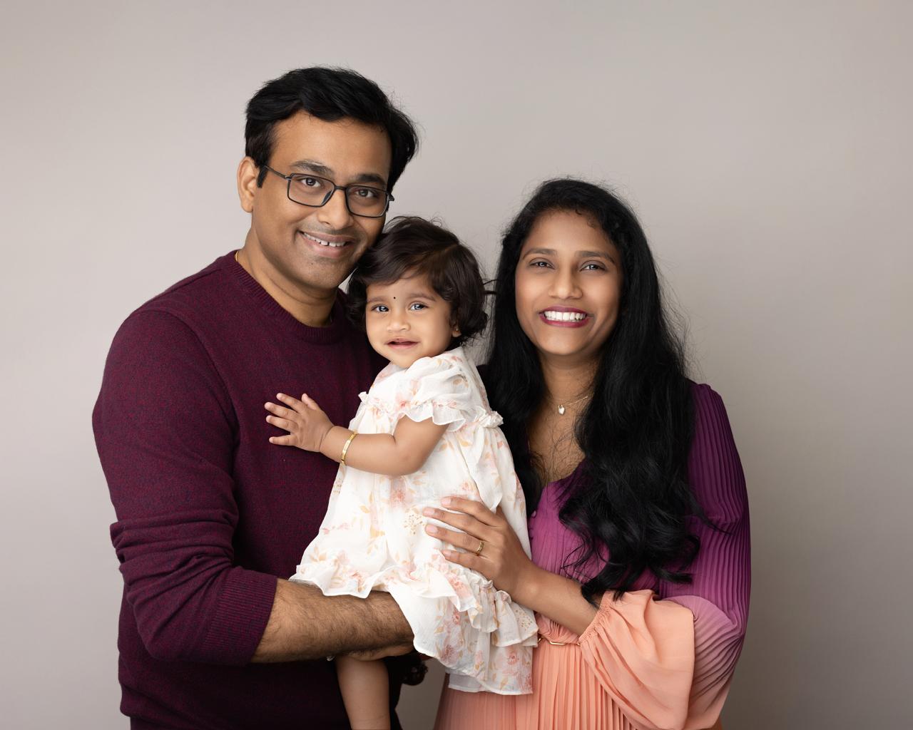 Smiling couple holding a young child dressed in a white floral dress against a plain background.