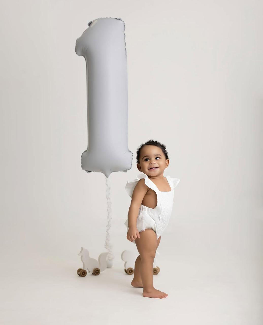 Smiling toddler in white outfit standing barefoot next to a silver balloon shaped like the number one and wooden toy animals on wheels.