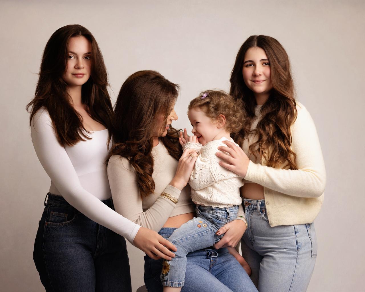 Four females with long hair, including a toddler sitting on a woman’s lap, smiling and interacting against a plain background.