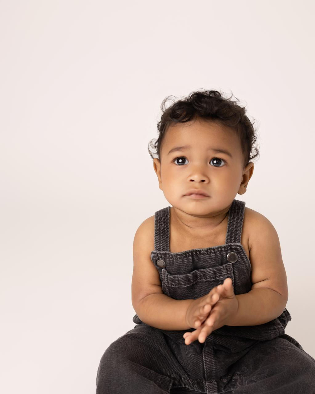 Young toddler with curly hair wearing dark overalls against a plain light background.