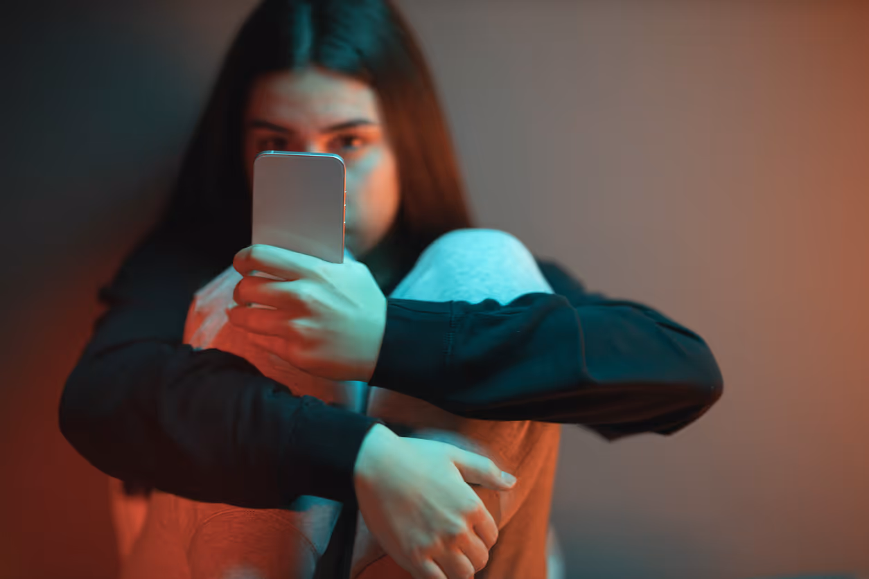 Meisje zit op de grond  met armen over haar benen en telefoon in haar hand