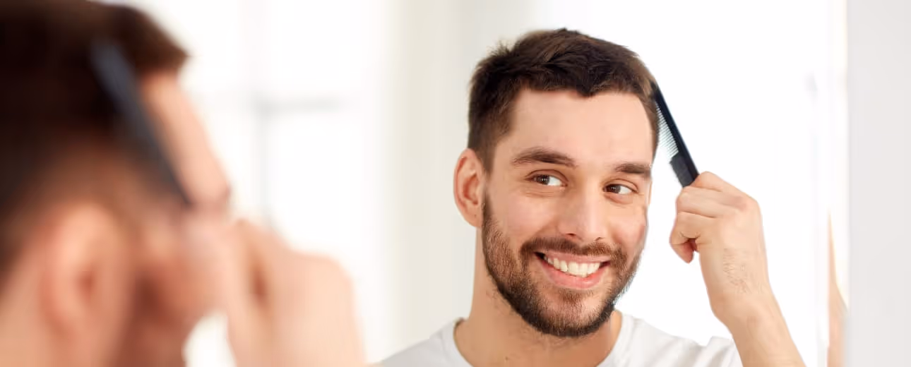 Smiling man combing his hair in front of a mirror, showcasing fuller hair after PRP hair restoration treatment.