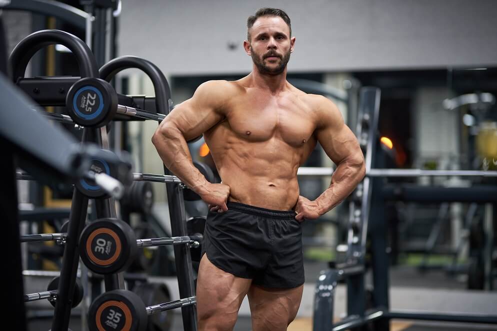 Muscular man posing in gym near weights, illustrating the long term benefits of testosterone replacement therapy (TRT).