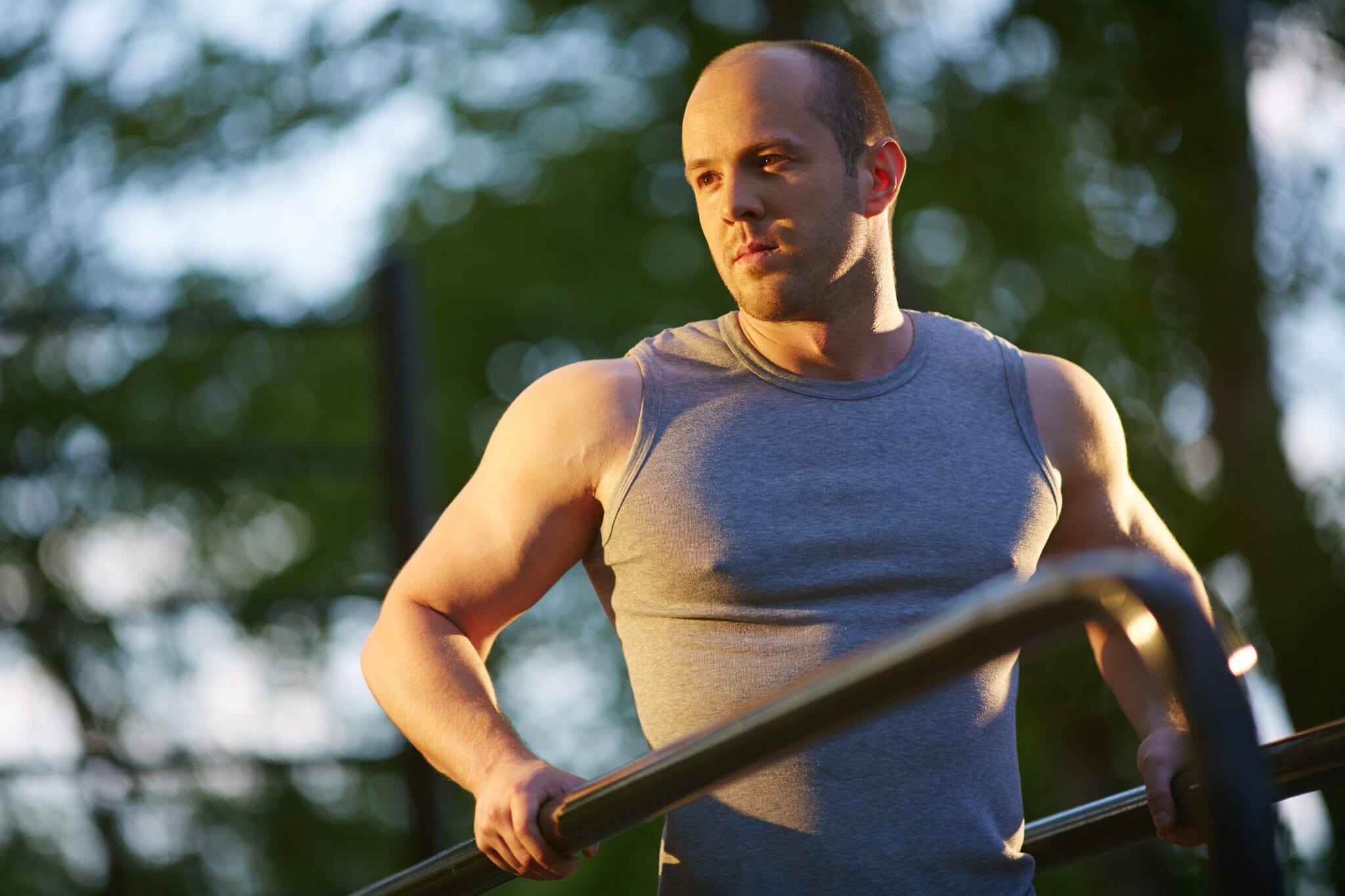 Fit man in a sleeveless shirt using parallel bars outdoors, symbolizing muscle health and physical performance benefits associated with peptide therapy.