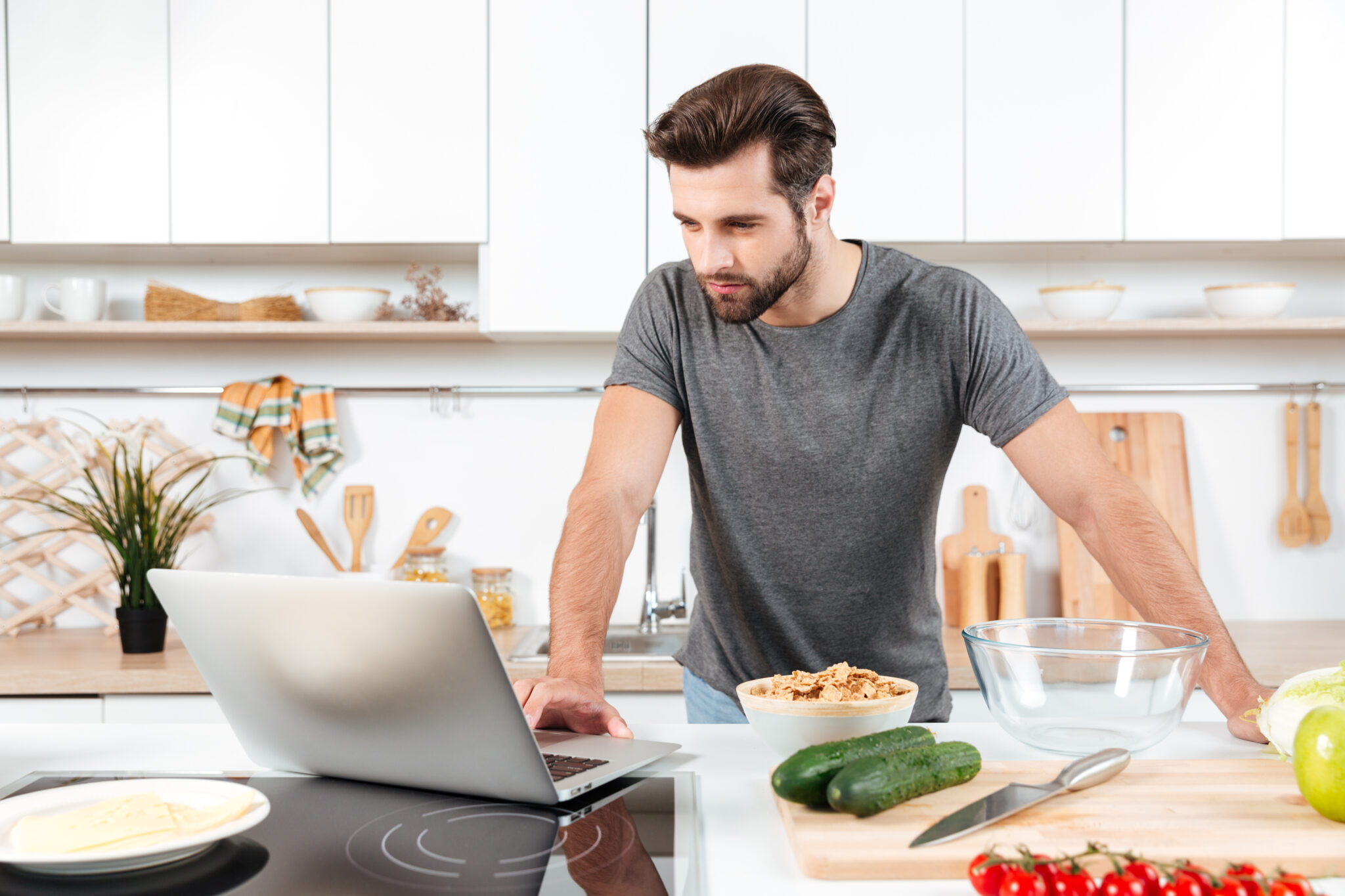 Man preparing a healthy meal in a modern kitchen while looking at a recipe on a laptop, with fresh vegetables and cereal on the counter.