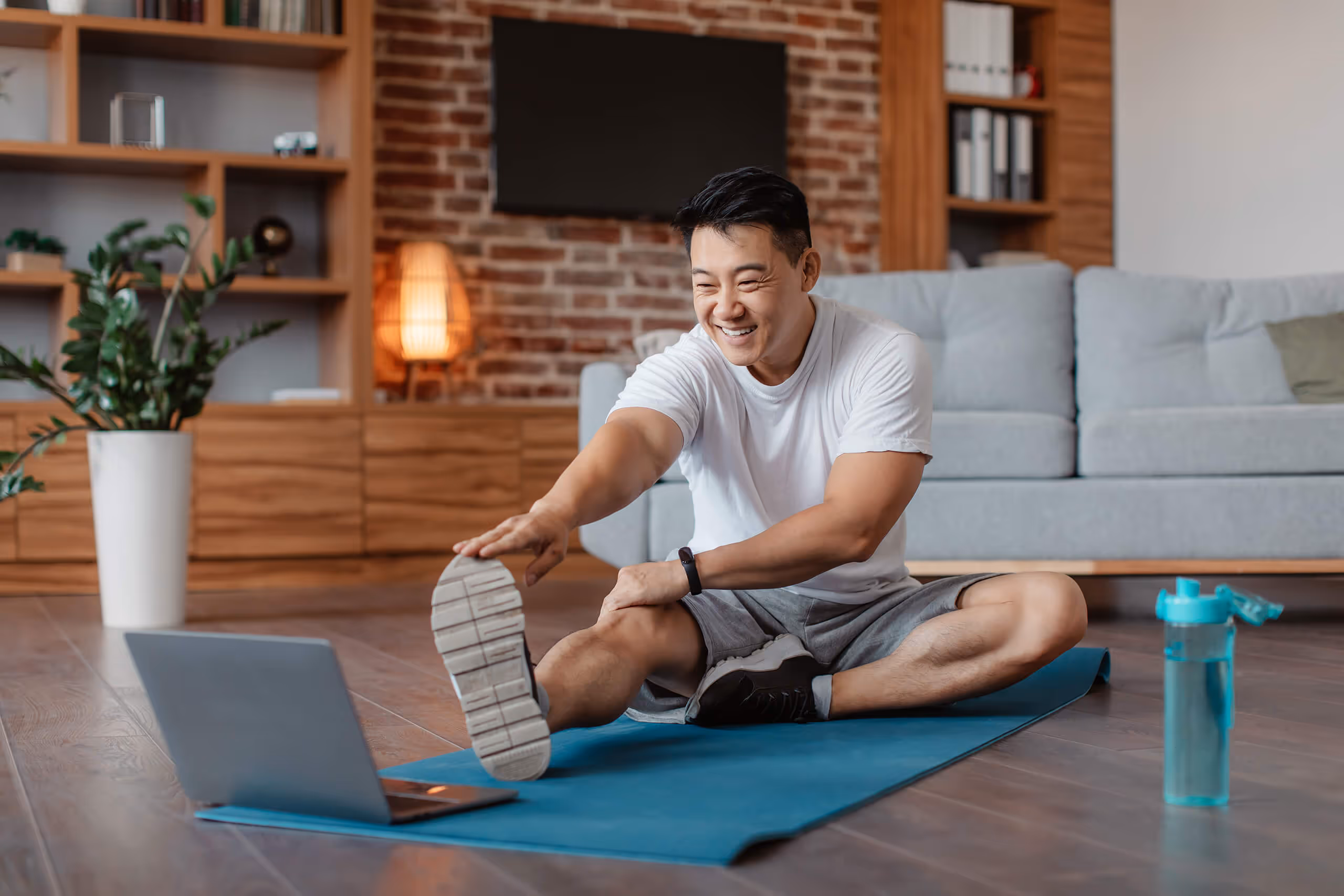Man smiling while stretching on a yoga mat during a virtual workout at home, promoting daily exercise as a habit for improved mental clarity