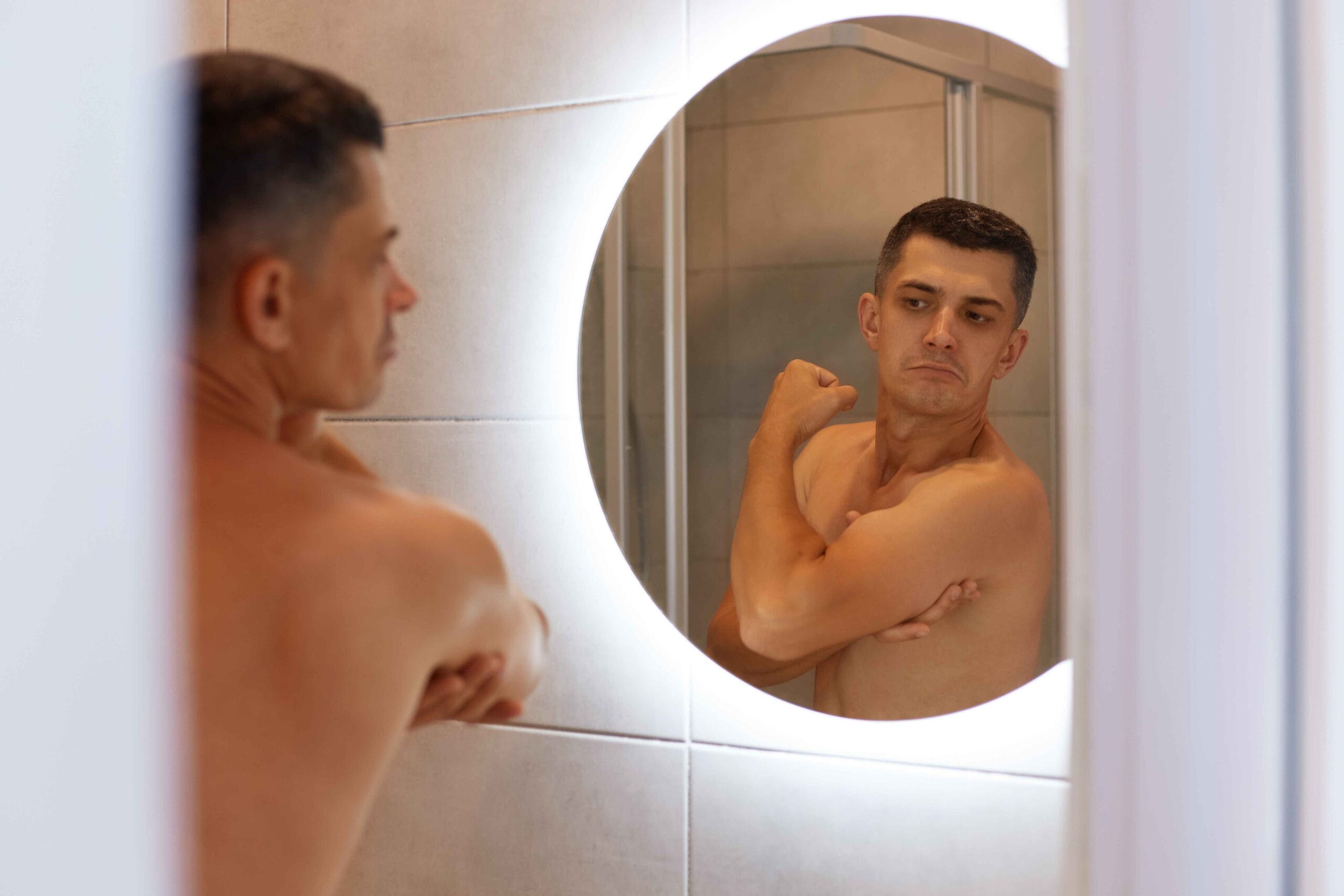 Man examining his muscles in a bathroom mirror, wondering whether low testosterone is affecting his ability to build muscle.