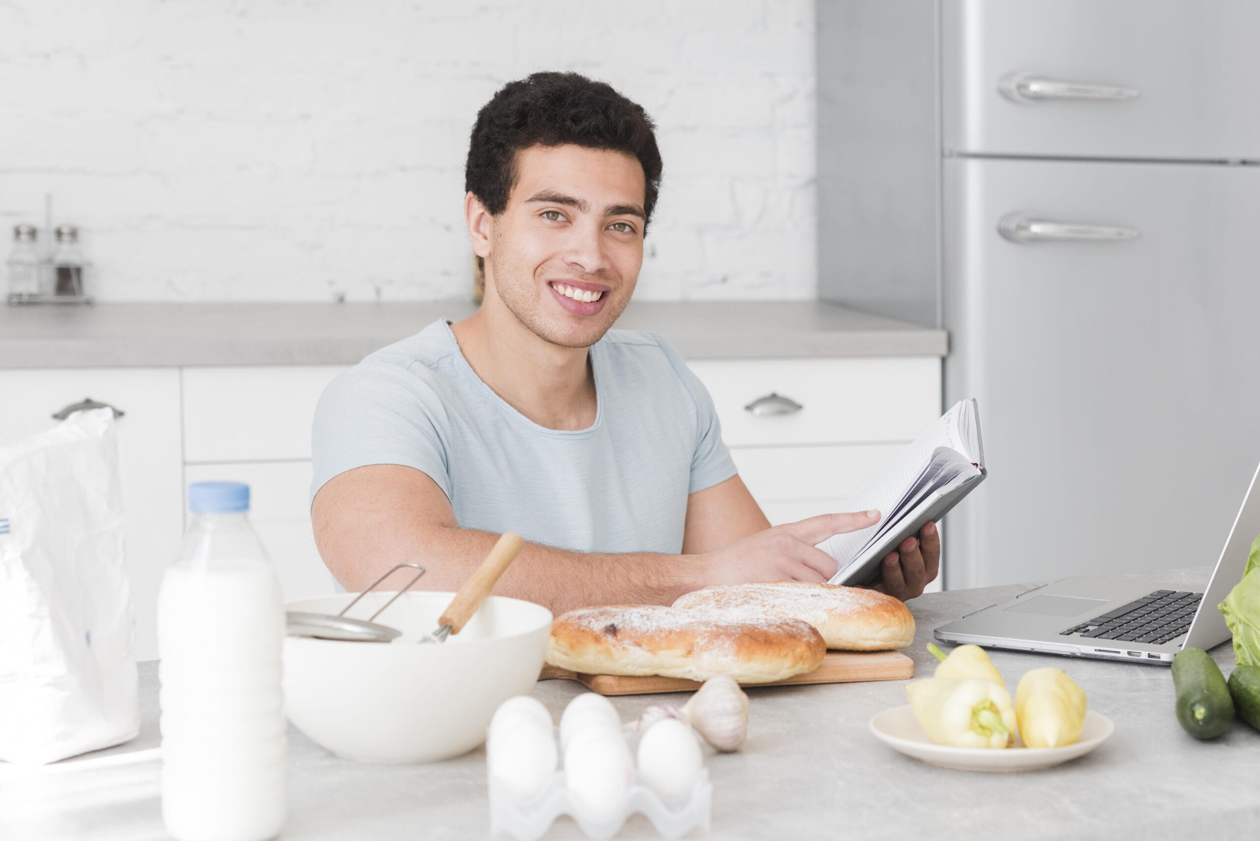 Smiling man reading a recipe book in the kitchen surrounded by bread, eggs, and vegetables, representing awareness of foods that may lower testosterone.