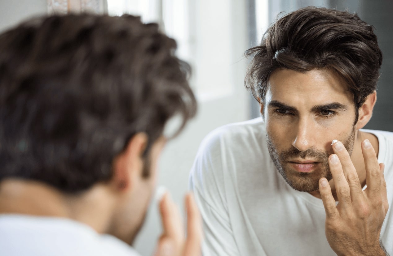 Man examining acne-prone skin in mirror, representing testosterone replacement therapy (TRT) and its impact on breakouts.