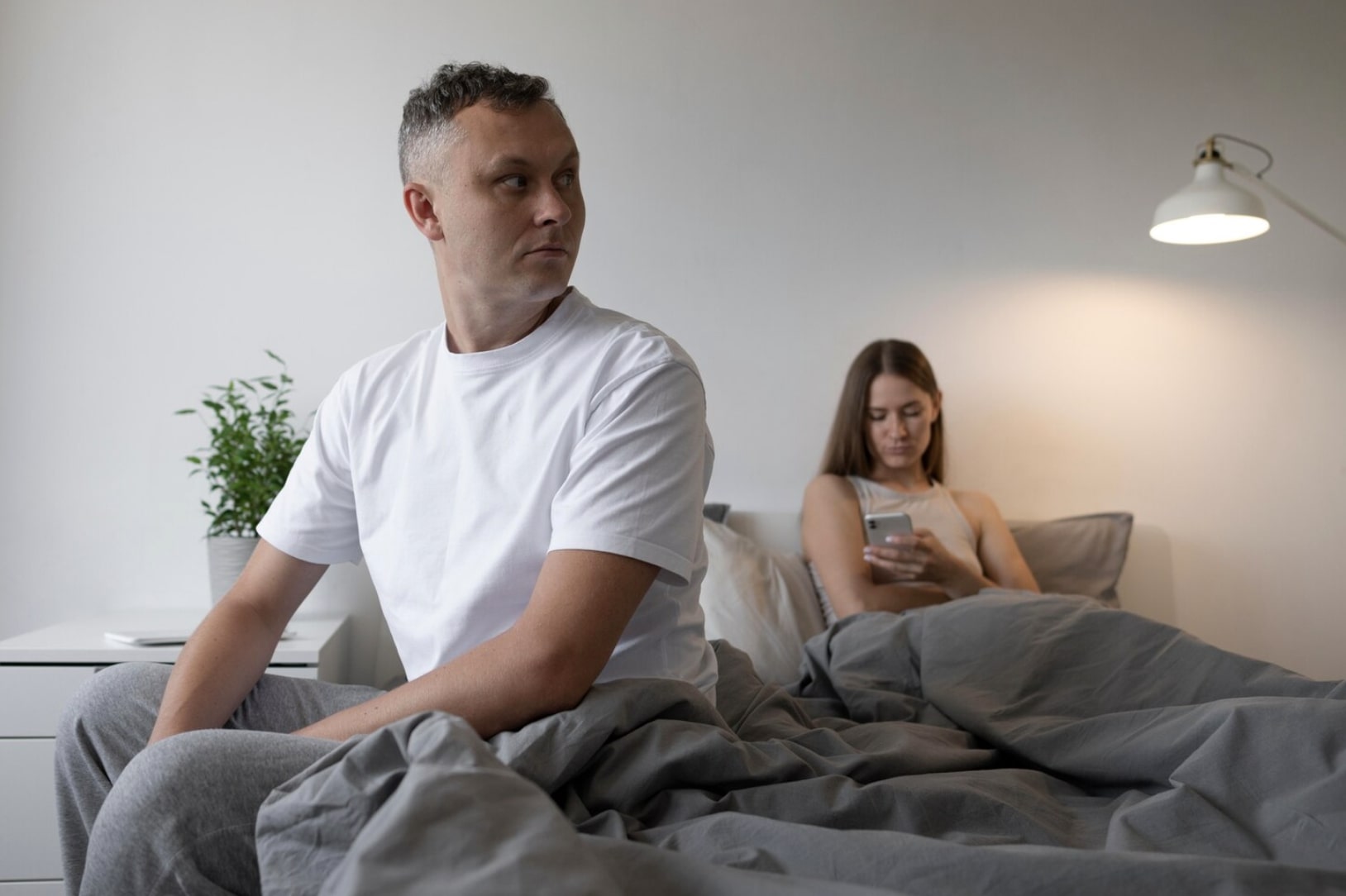 A concerned man sitting on the edge of the bed with his partner in the background, representing the emotional impact and uncertainty of temporary erectile dysfunction.