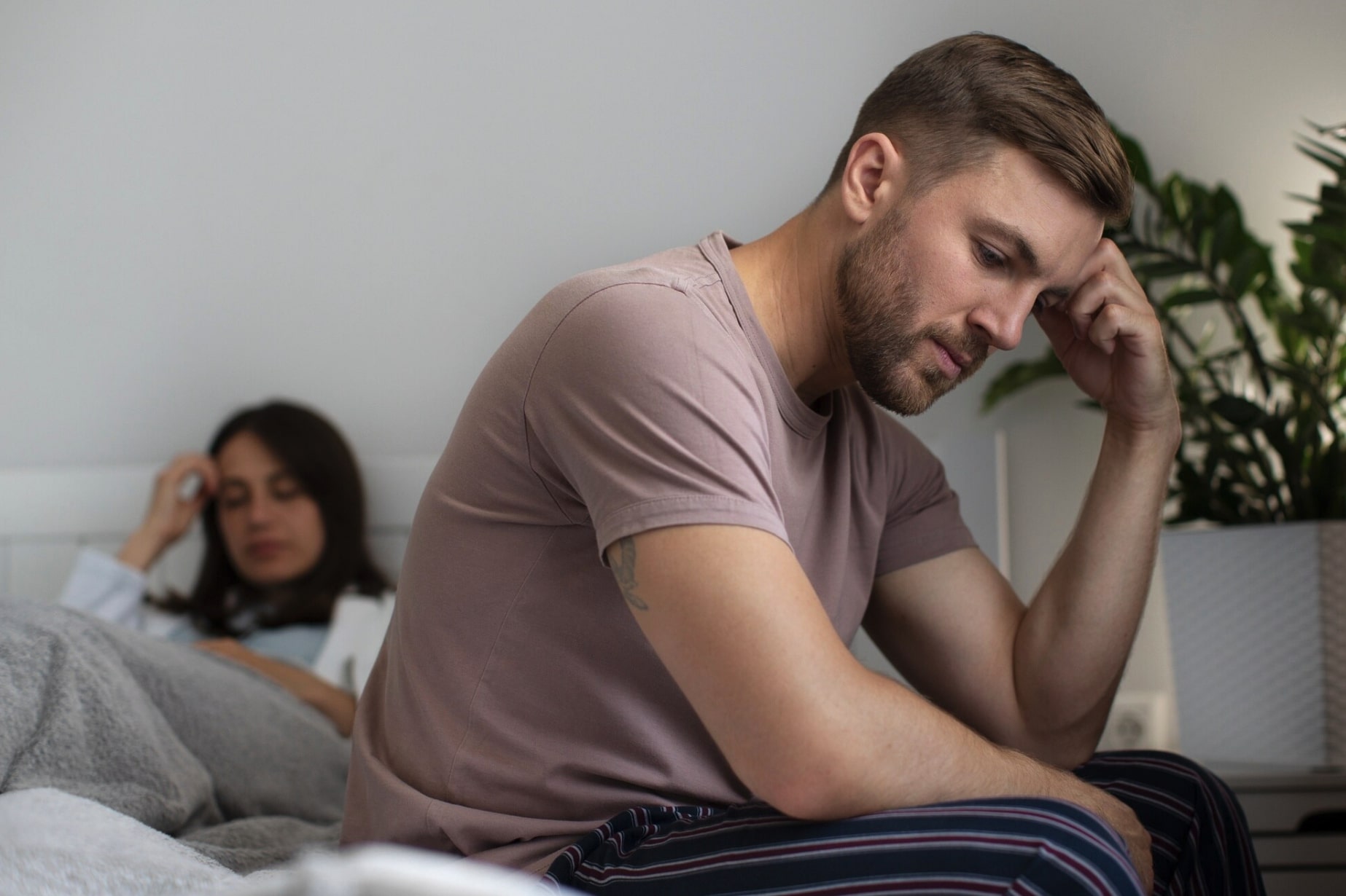 Man and woman sitting apart on a bed looking concerned, illustrating relationship stress related to erectile dysfunction and medication side effects.