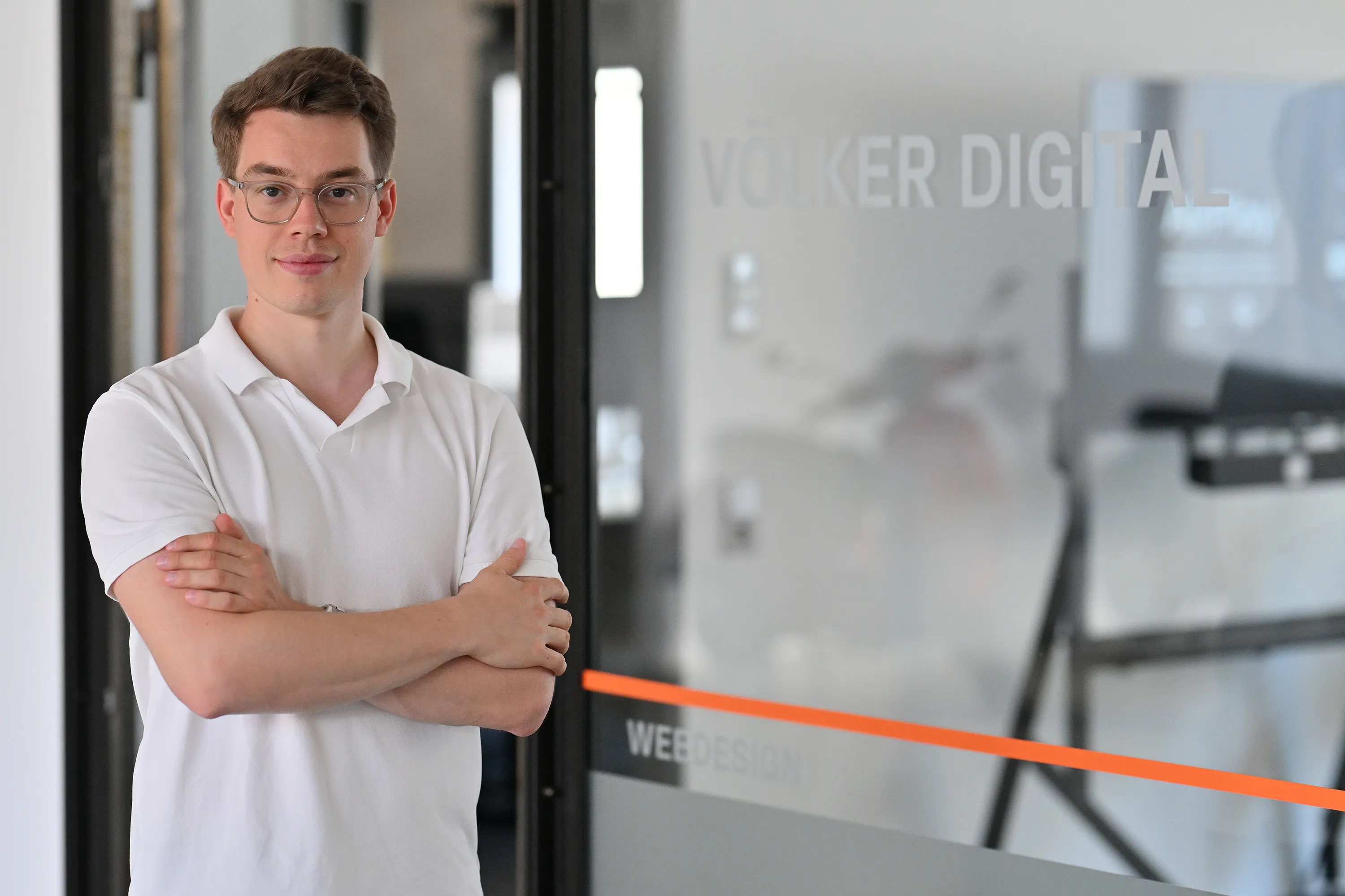 Junger Mann mit Brille und weißem Poloshirt steht mit verschränkten Armen vor einer Glaswand mit der Aufschrift ‚VÖLKER DIGITAL WEB DESIGN‘.