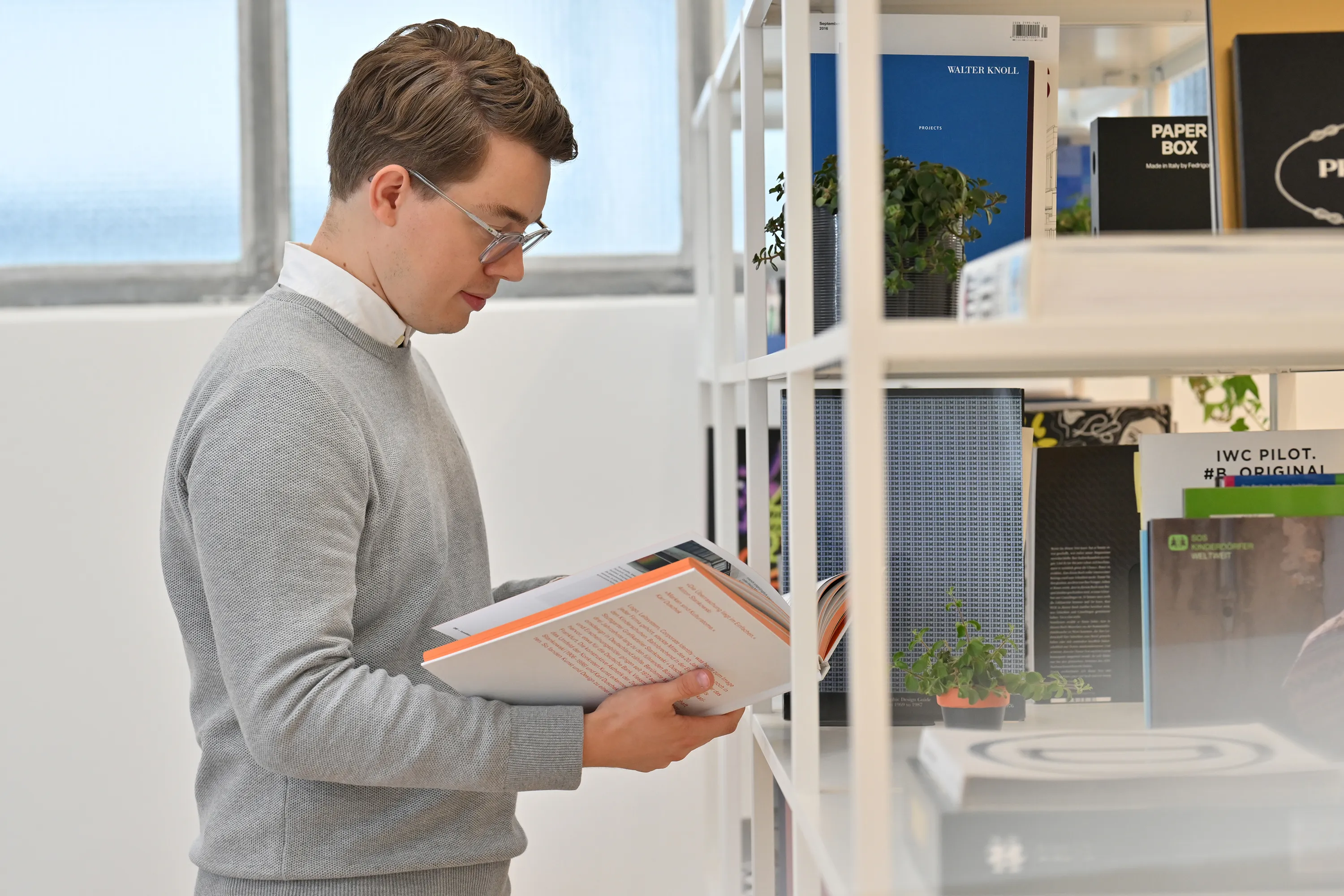Mann mit Brille und grauem Pullover liest in einem offenen Buch vor einem Regal mit Pflanzen und Büchern.