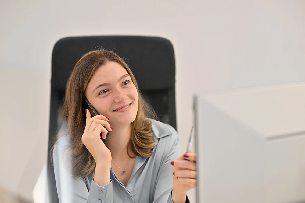 Junge Frau sitzt lächelnd auf einem Bürostuhl, telefoniert und hält einen Stift vor einem Computerbildschirm.
