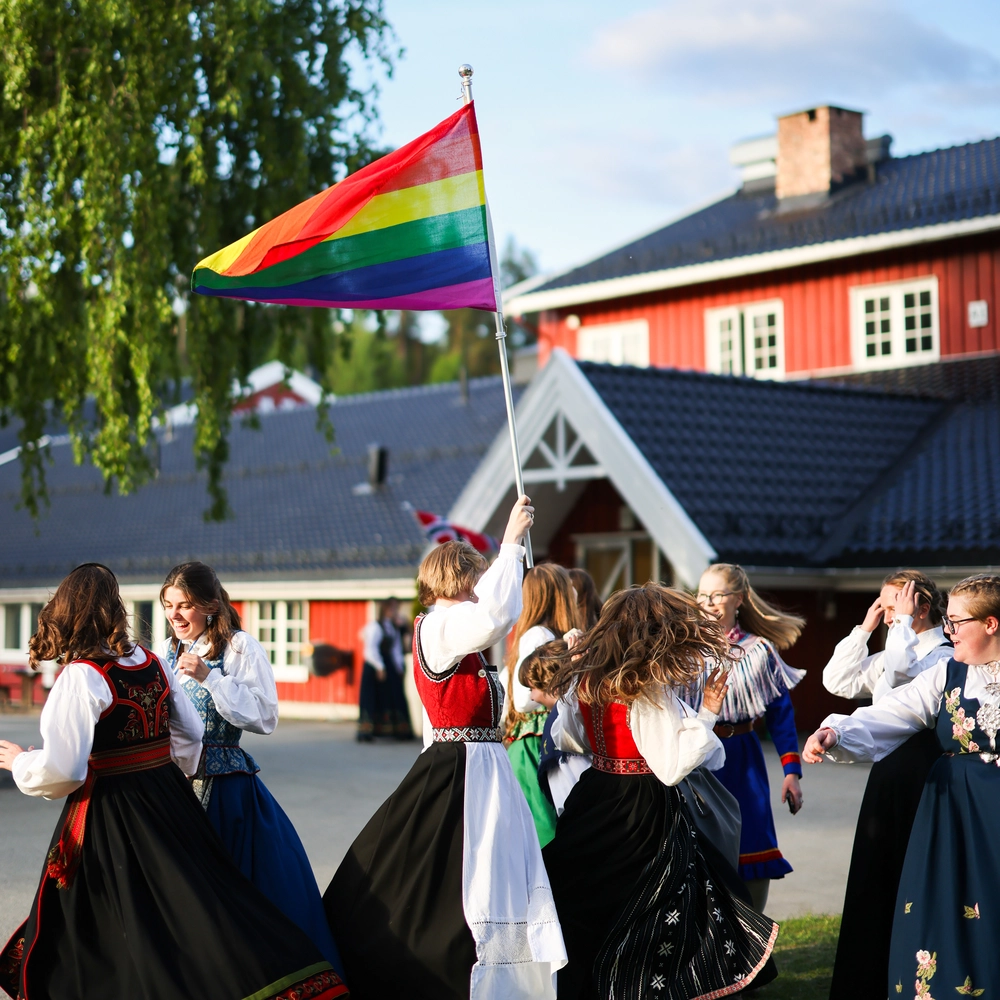 Elever ved Ringerike Folkehøgskole danser i bunad mens de vifter med regnbueflagget, et bilde som symboliserer mangfold, inkludering og glede i skolefellesskapet.