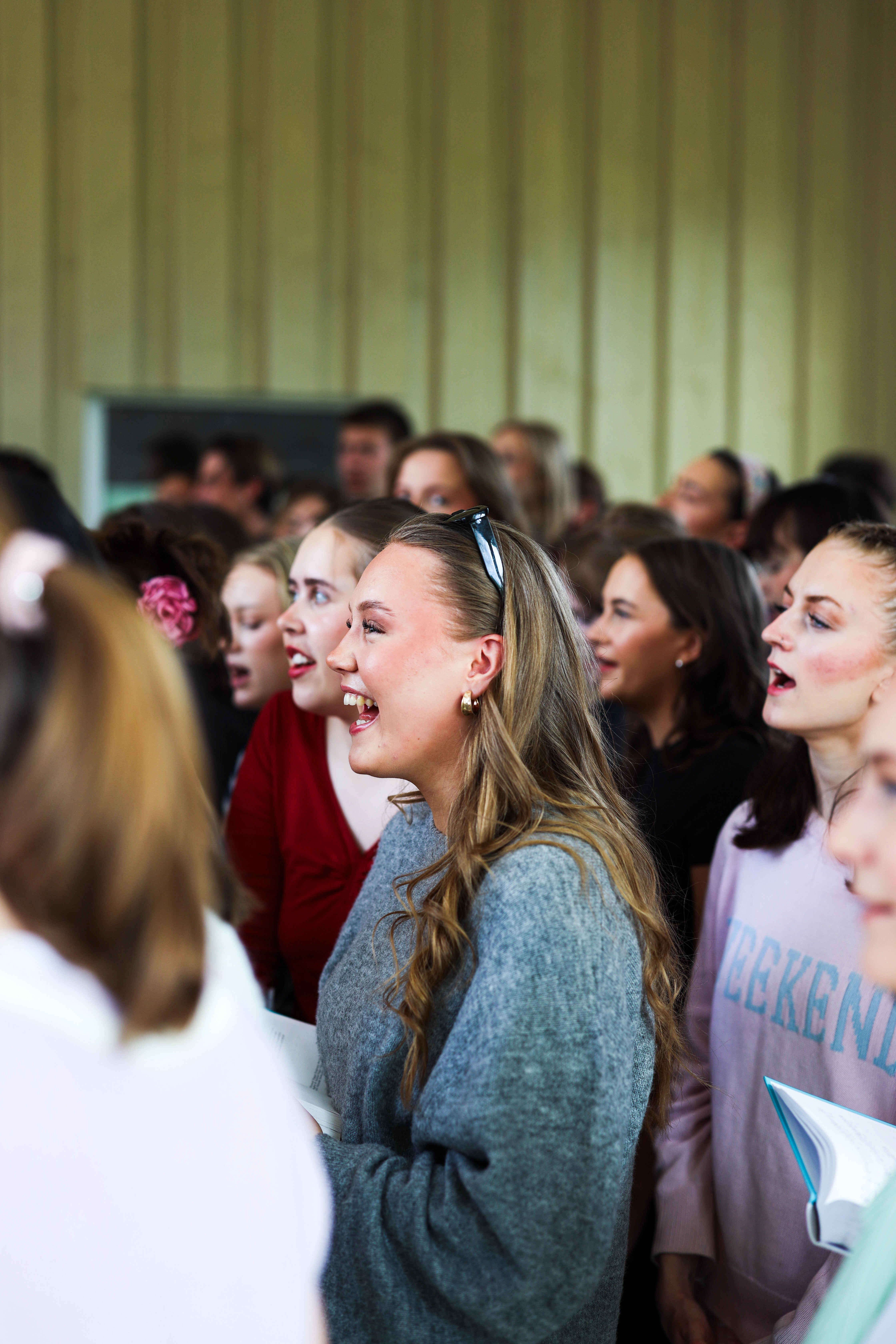 Skolekoret synger sammen innendørs med smil og engasjement, et bilde som fanger fellesskap, glede og samhold på Ringerike Folkehøgskole.
