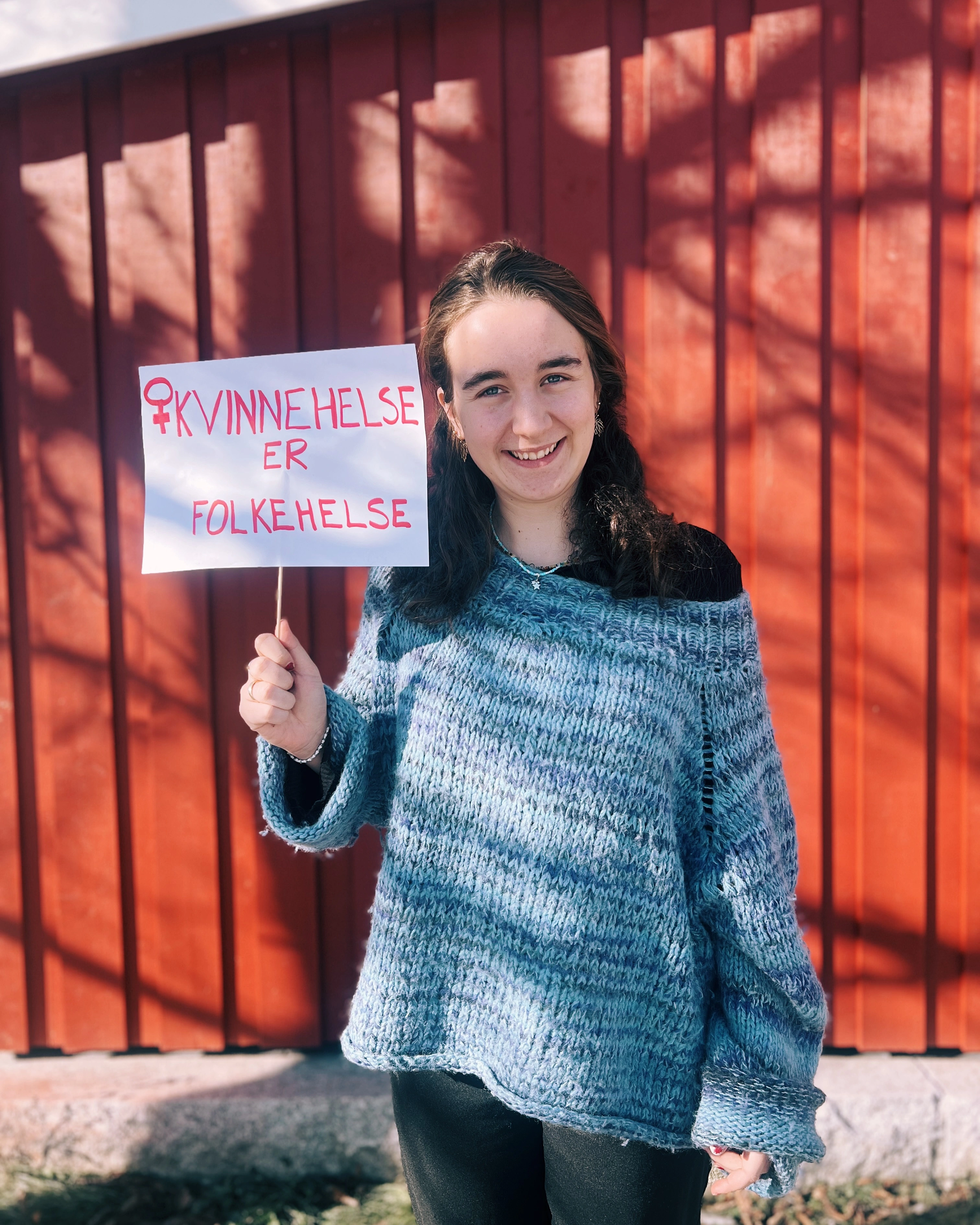 En elev smiler foran en rød vegg og holder et skilt med teksten «Kvinnehelse er folkehelse», et bilde som symboliserer engasjement og solidaritet.