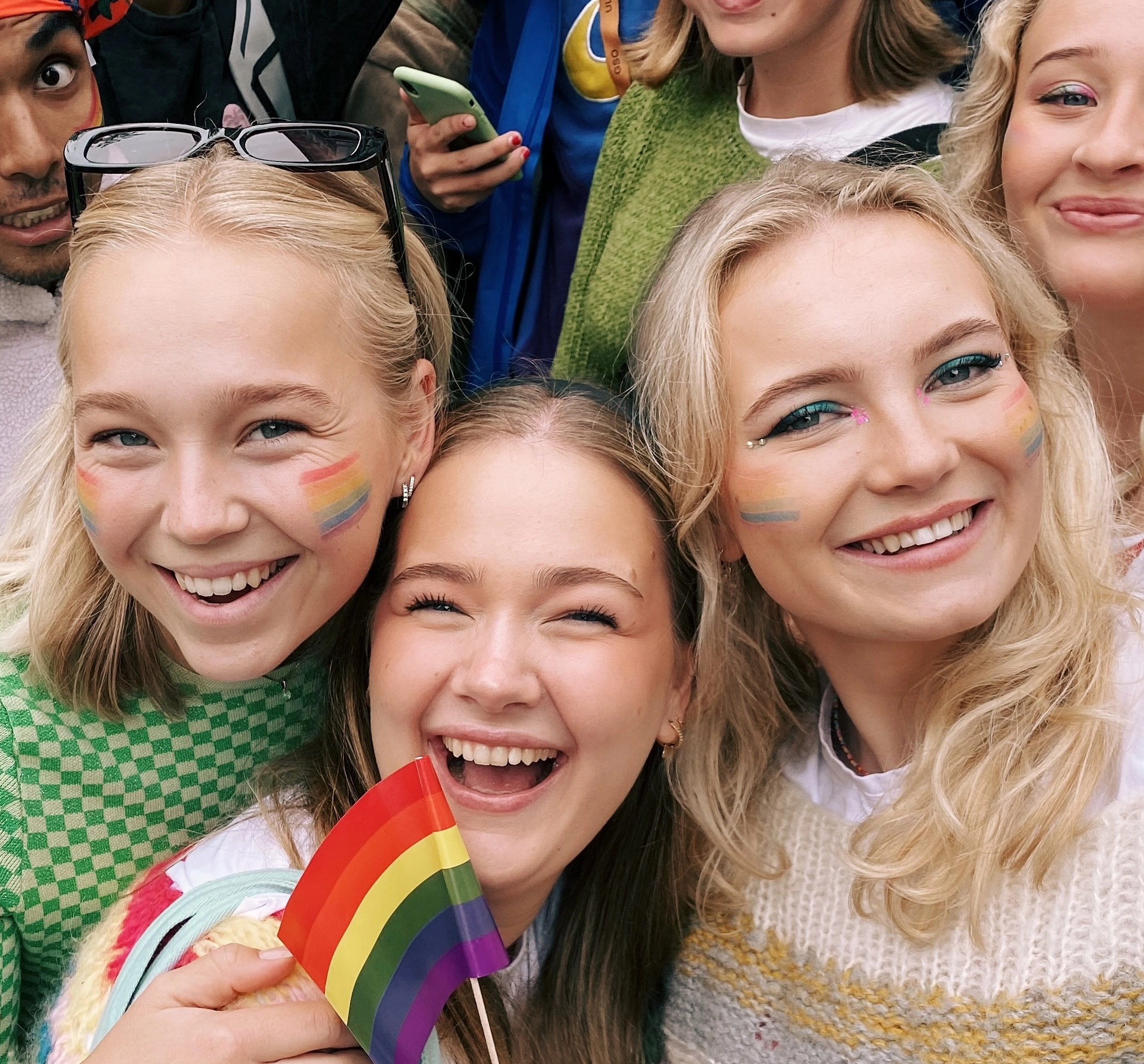 Tre elever smiler med regnbuemaling på kinnene og holder et prideflagg, et bilde som uttrykker inkludering, mangfold og fellesskap ved Ringerike Folkehøgskole.