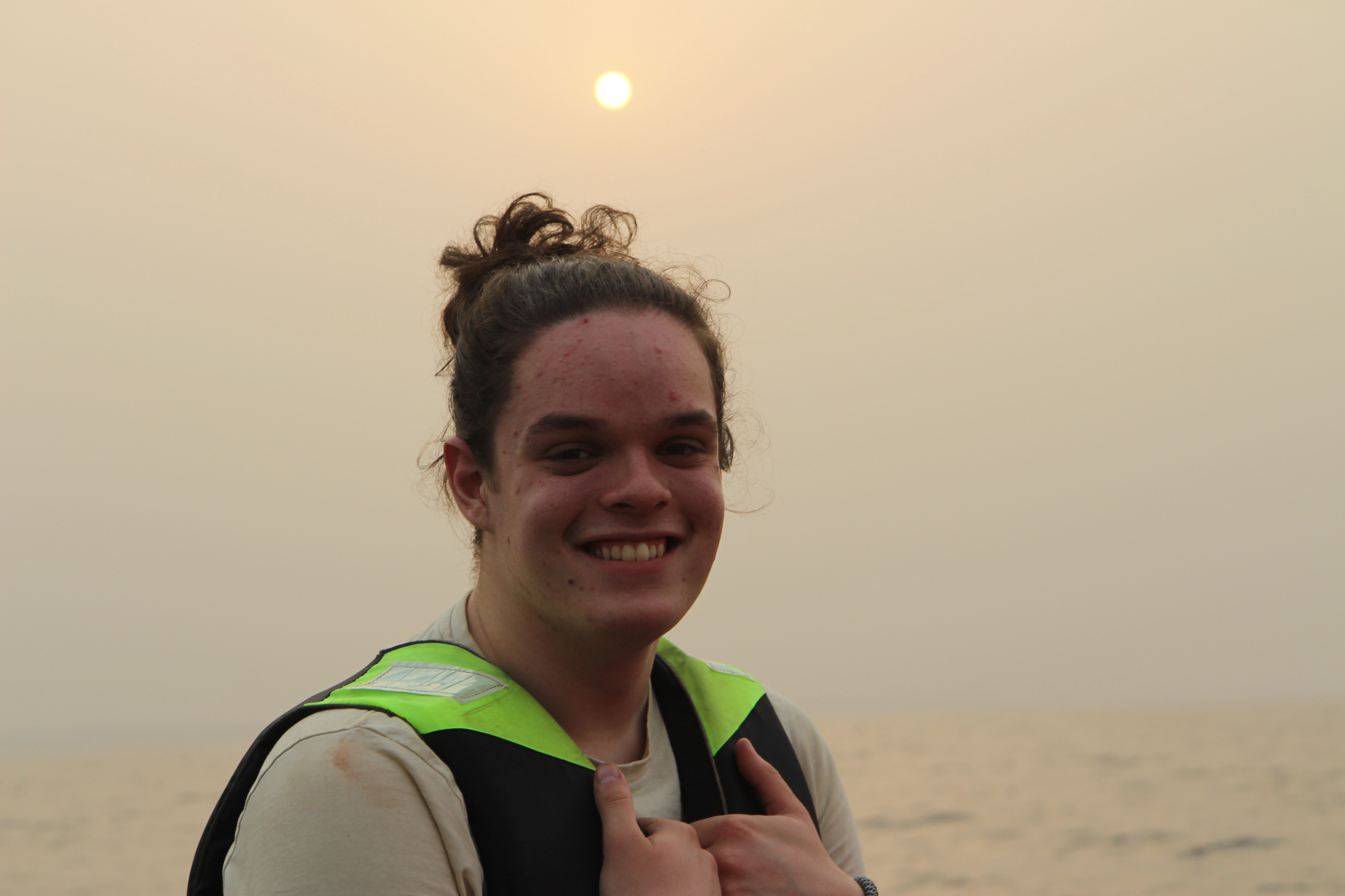 Elev fra linjen UT på Ringerike Folkehøgskole smiler ved sjøen under solnedgang i Ghana, et bilde som viser eventyr, reiselyst og friluftsliv.