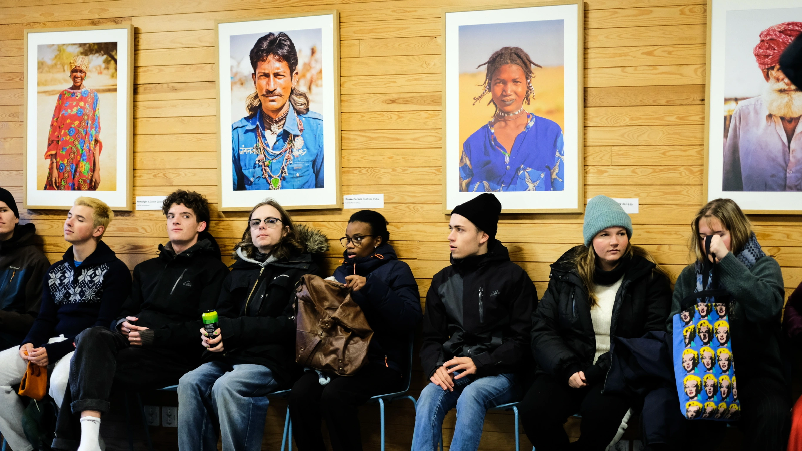 Elever ved Ringerike Folkehøgskole sitter foran veggen med portrettfotografier fra ulike kulturer, et bilde som viser refleksjon, nysgjerrighet og globalt perspektiv.