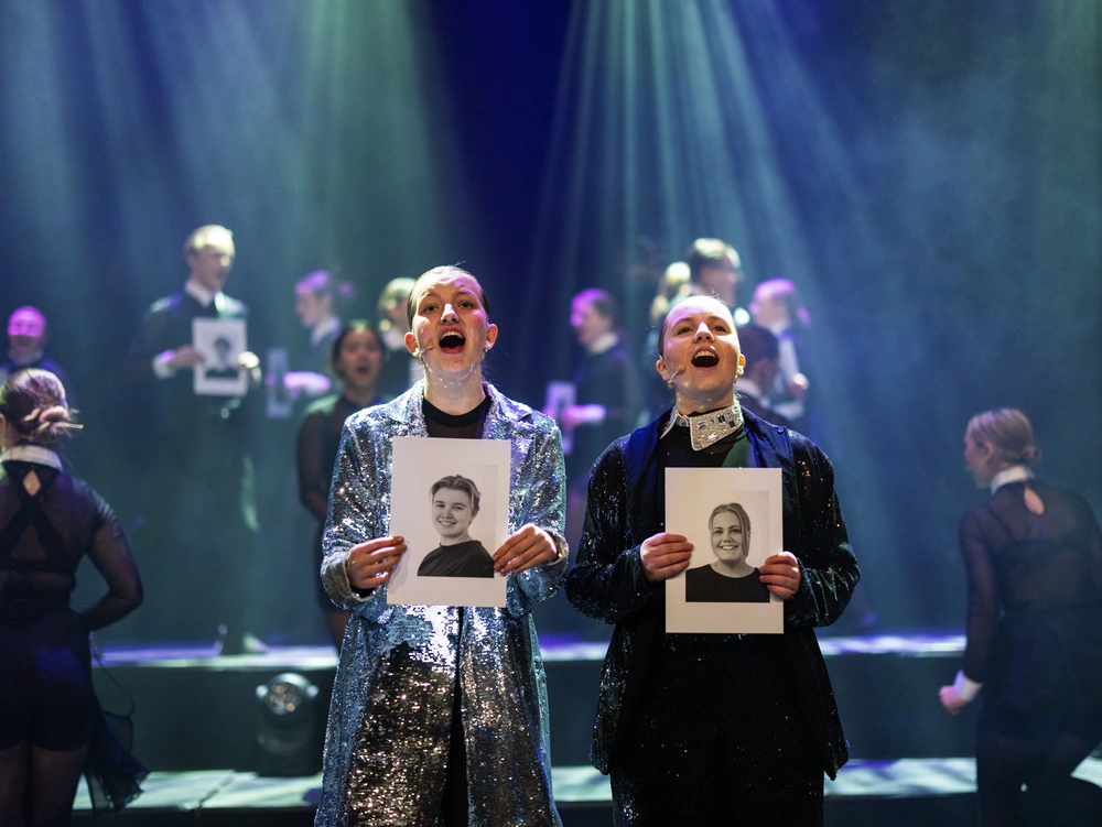 Elever fra linjen Musikal på Ringerike Folkehøgskole synger på scenen under forestilling med lys og kostymer, et bilde som viser engasjement og sceneglede.