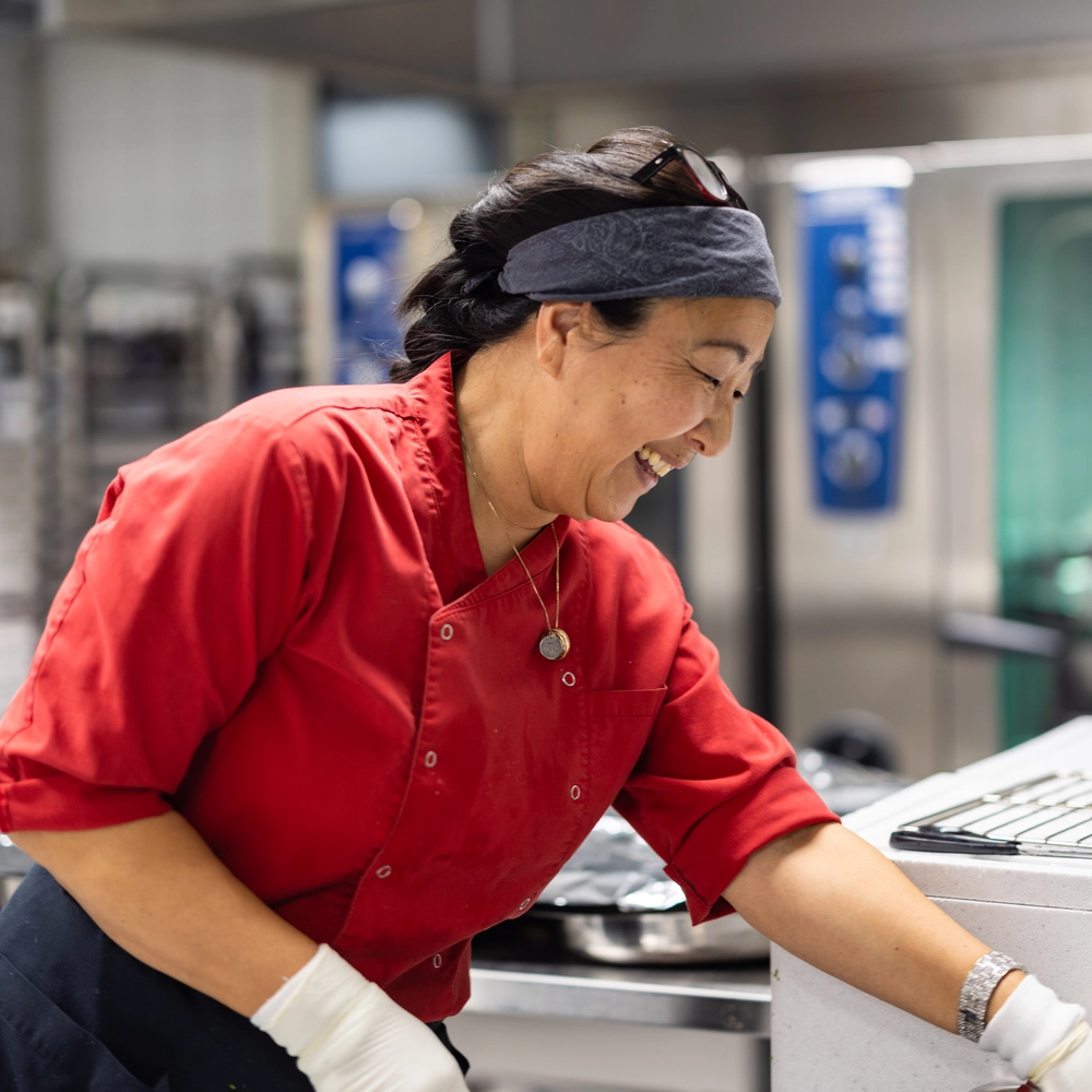 Kjøkkenansatt smiler mens hun lager mat på Ringerike Folkehøgskole, et bilde som viser trivsel og god mat laget med omtanke.