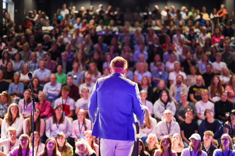 Foredragsholder står på scenen foran fullsatt sal under åpning eller arrangement på Ringerike Folkehøgskole, et bilde som viser fellesskap og engasjement.