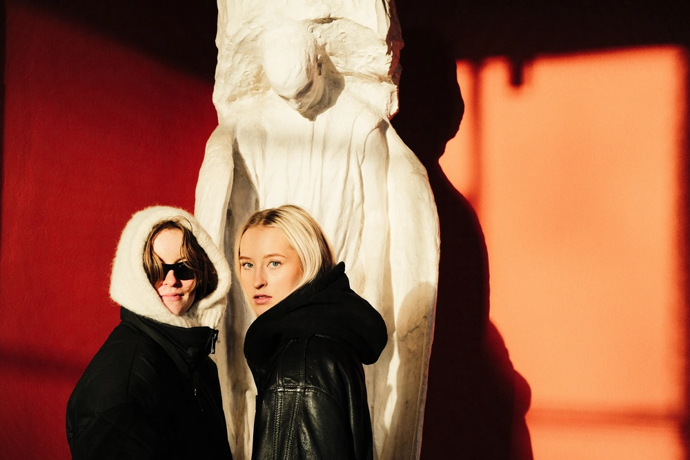 To elever fra Ringerike Folkehøgskole poserer foran en hvit skulptur mot rød vegg, som del av skolens kreative foto- og kunstprosjekt.