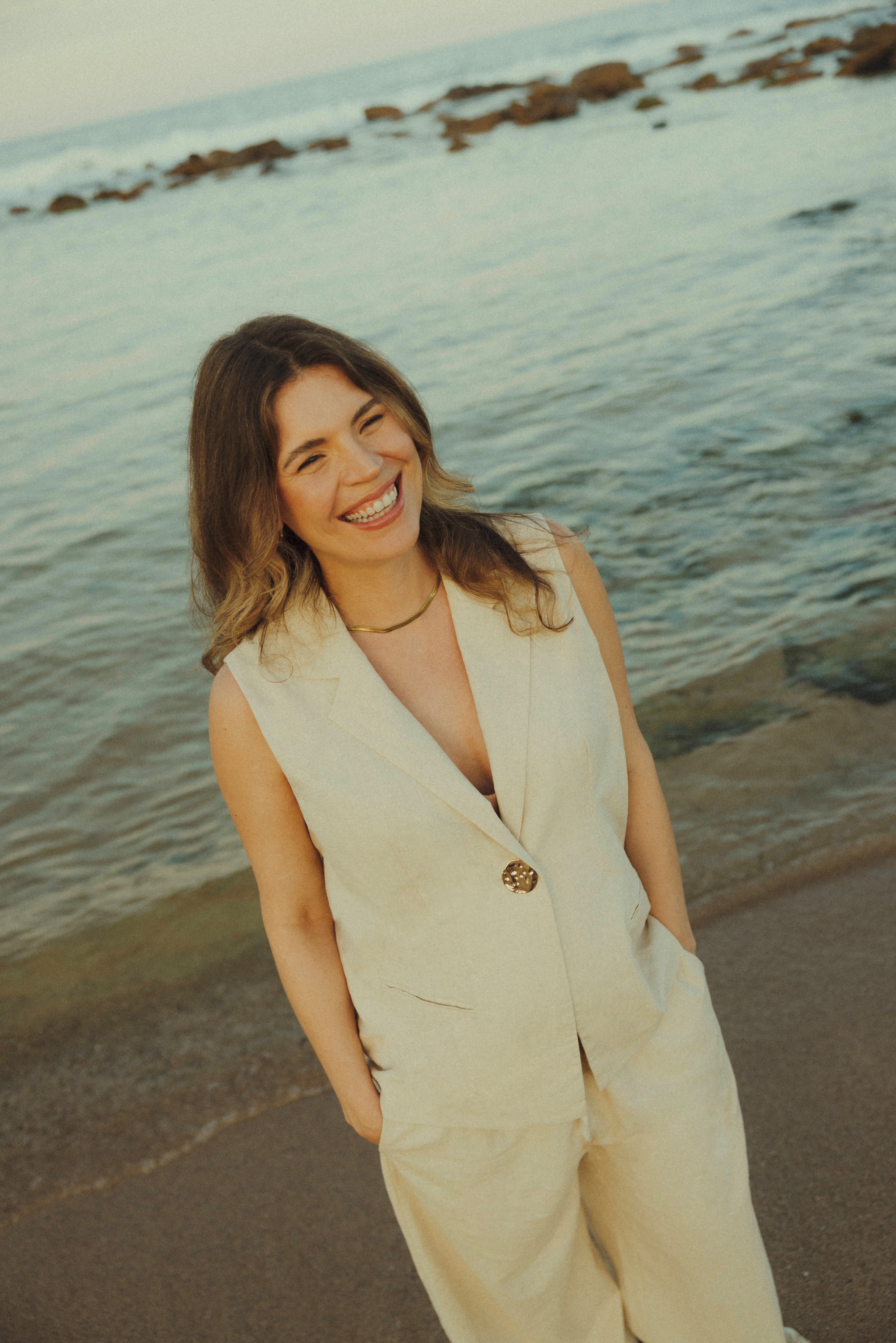 Smiling Mi'Ora Founder wearing a beige vest and pants standing on a beach with ocean waves and rocks in the background.