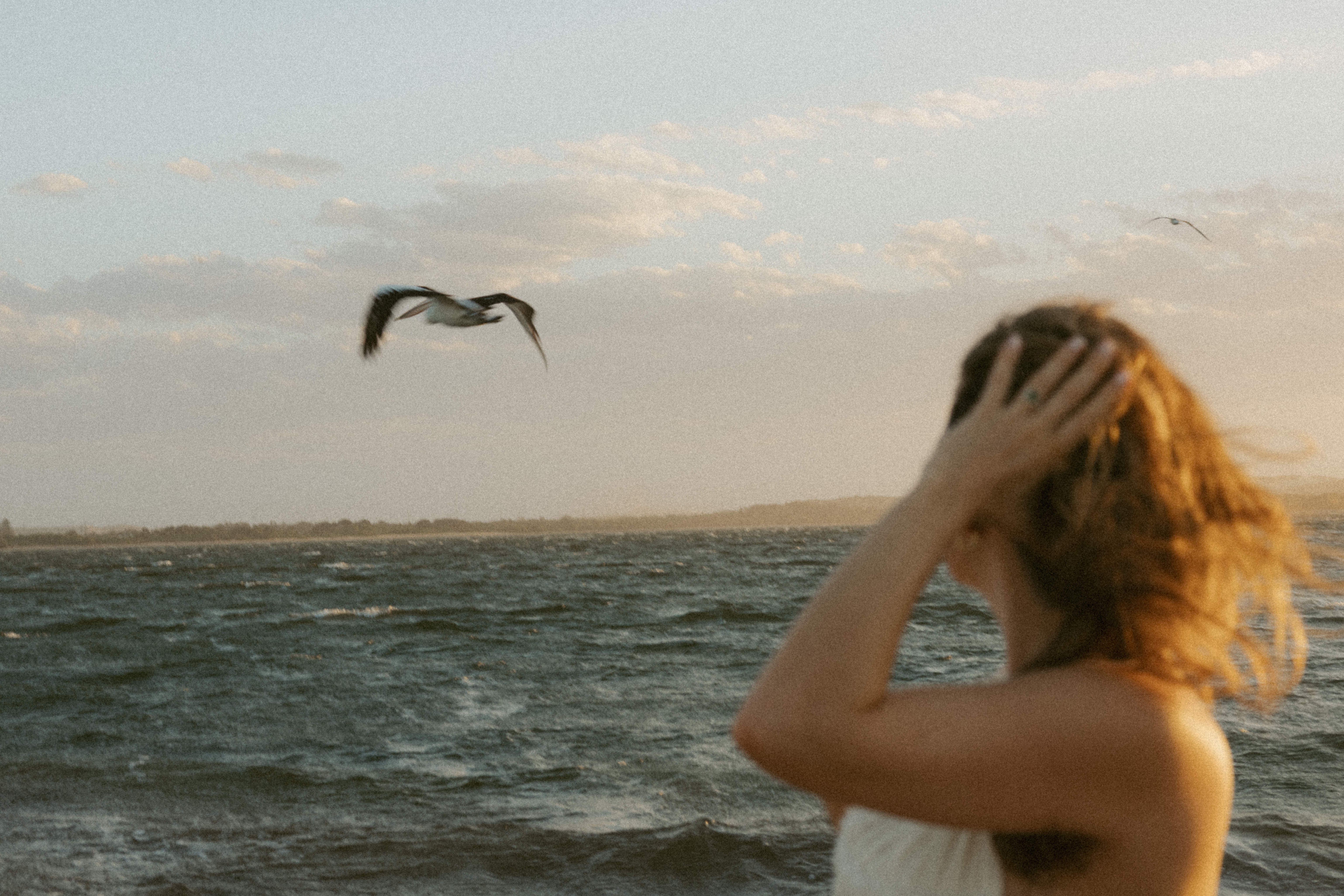 Mi'Ora Founder with windblown hair touching her head while looking at pelican flying over a choppy sea at sunset.