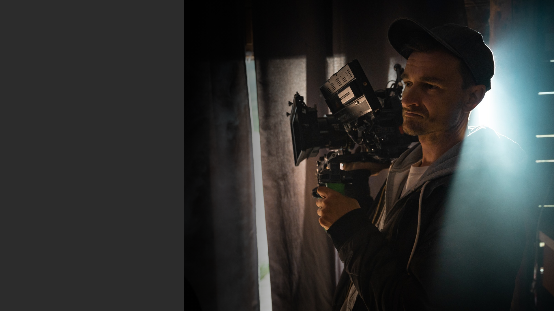 Man operating a professional video camera indoors with a focused expression and backlighting behind him.