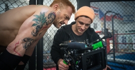 Two men reviewing footage on a professional video camera inside a gym with a caged fighting ring in the background.