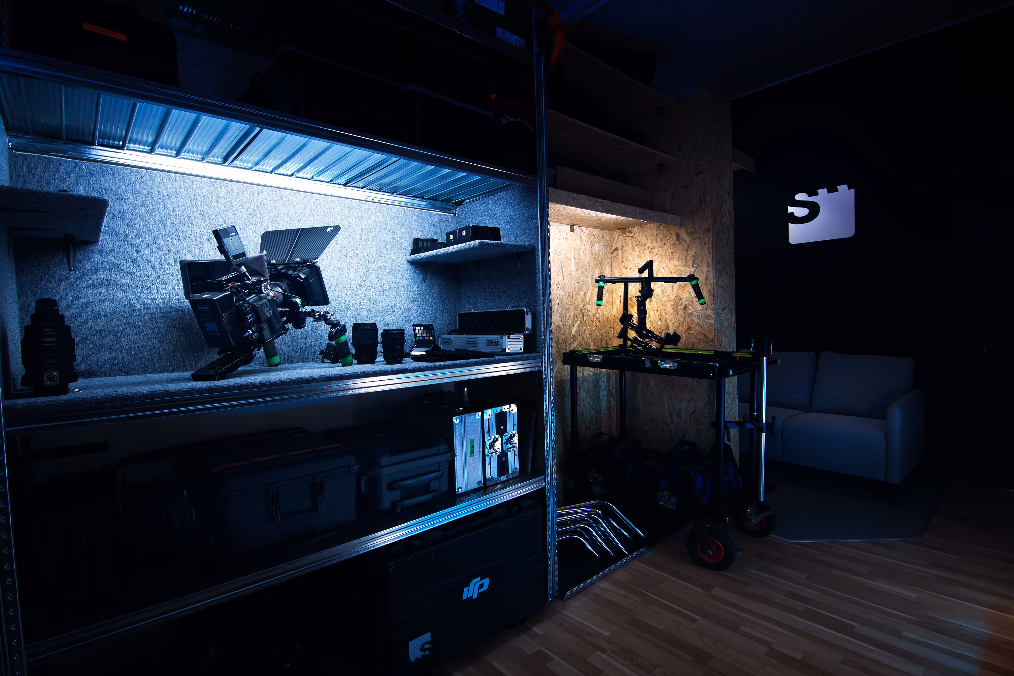Dimly lit room with shelves holding professional camera equipment and a cart with a camera stabilizer next to a white couch.