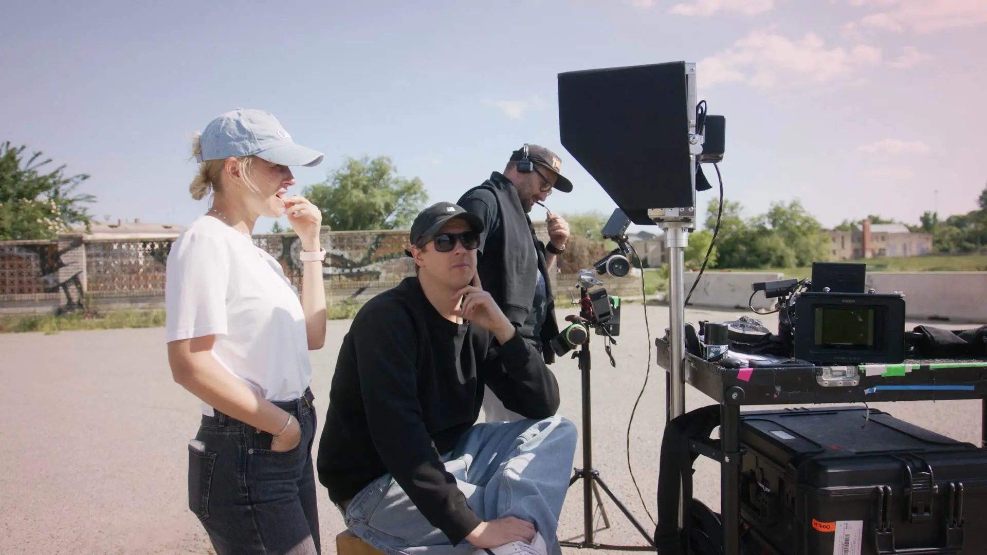 Tři lidé stojí a sedí venku vedle filmového vybavení za slunečného dne.