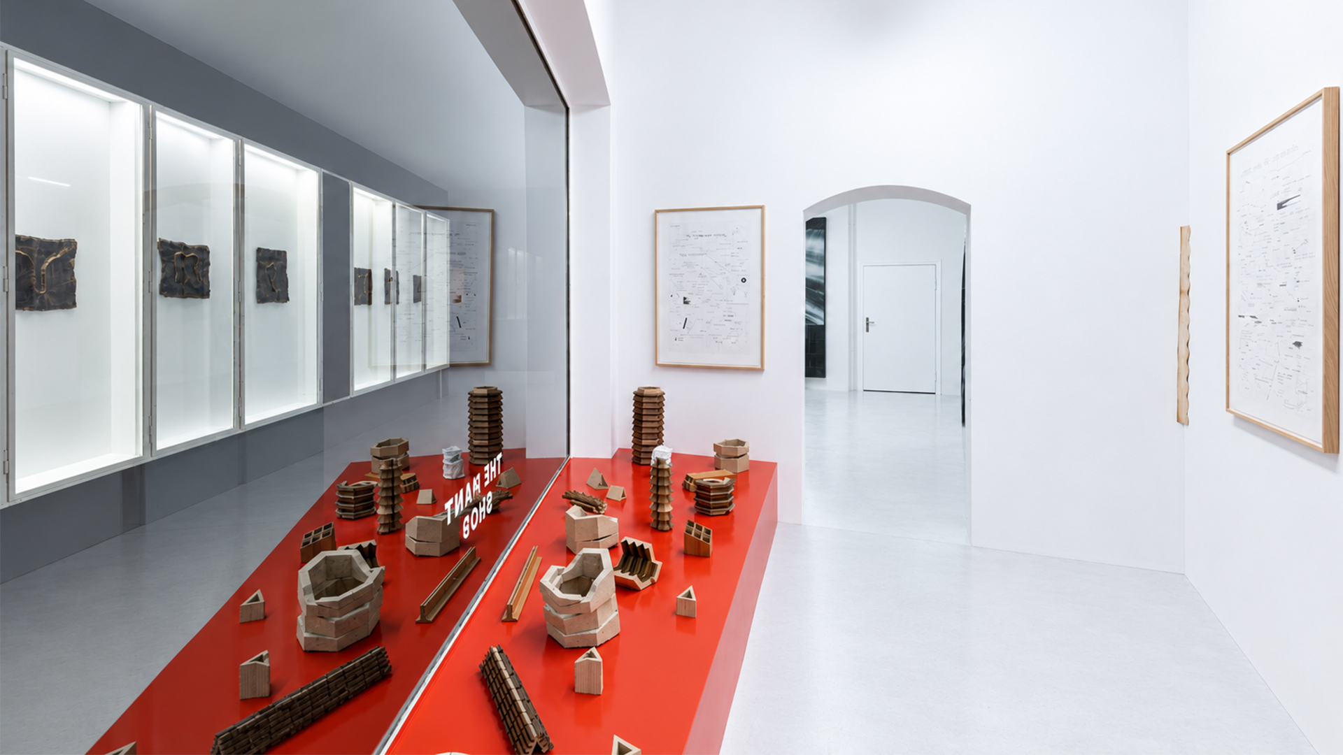 Inside Paint Shop gallery, Hasenheide 12, Berlin: sculptures on a long red display table, framed works on white walls and lit vitrines