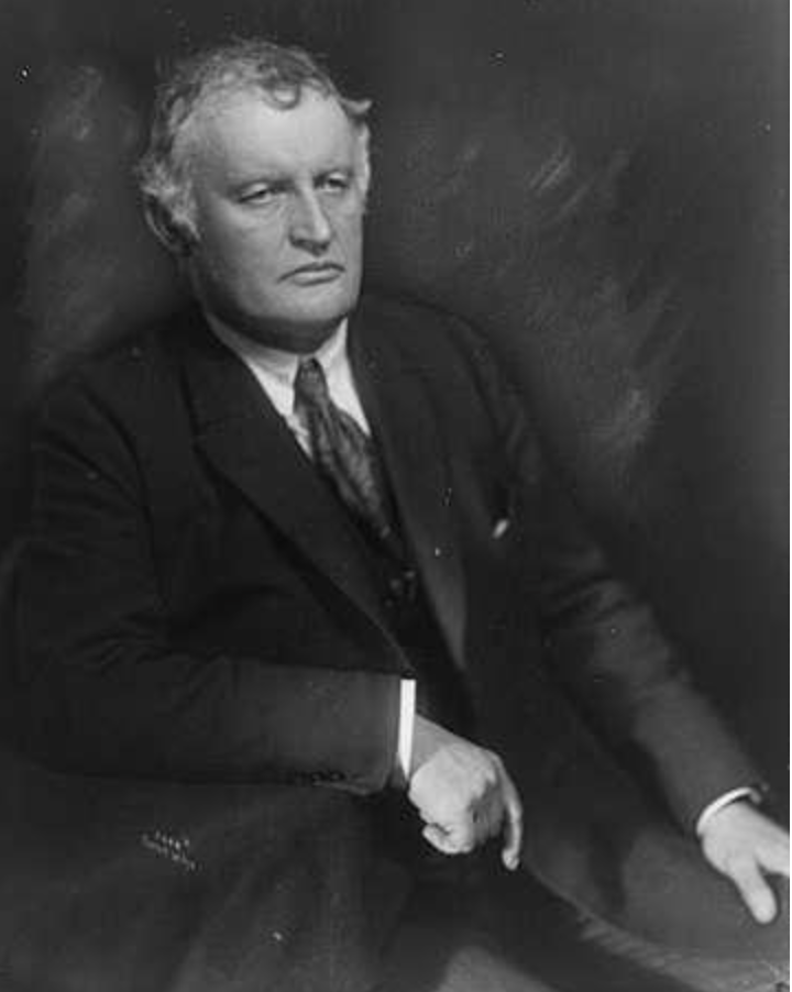 Edvard Munch by Anders Beer Wilse (1921) - Black and white portrait of Edvard Munch in a suit in 1921, seated against a dark backdrop. His expression is serious, with a focused and introspective look.