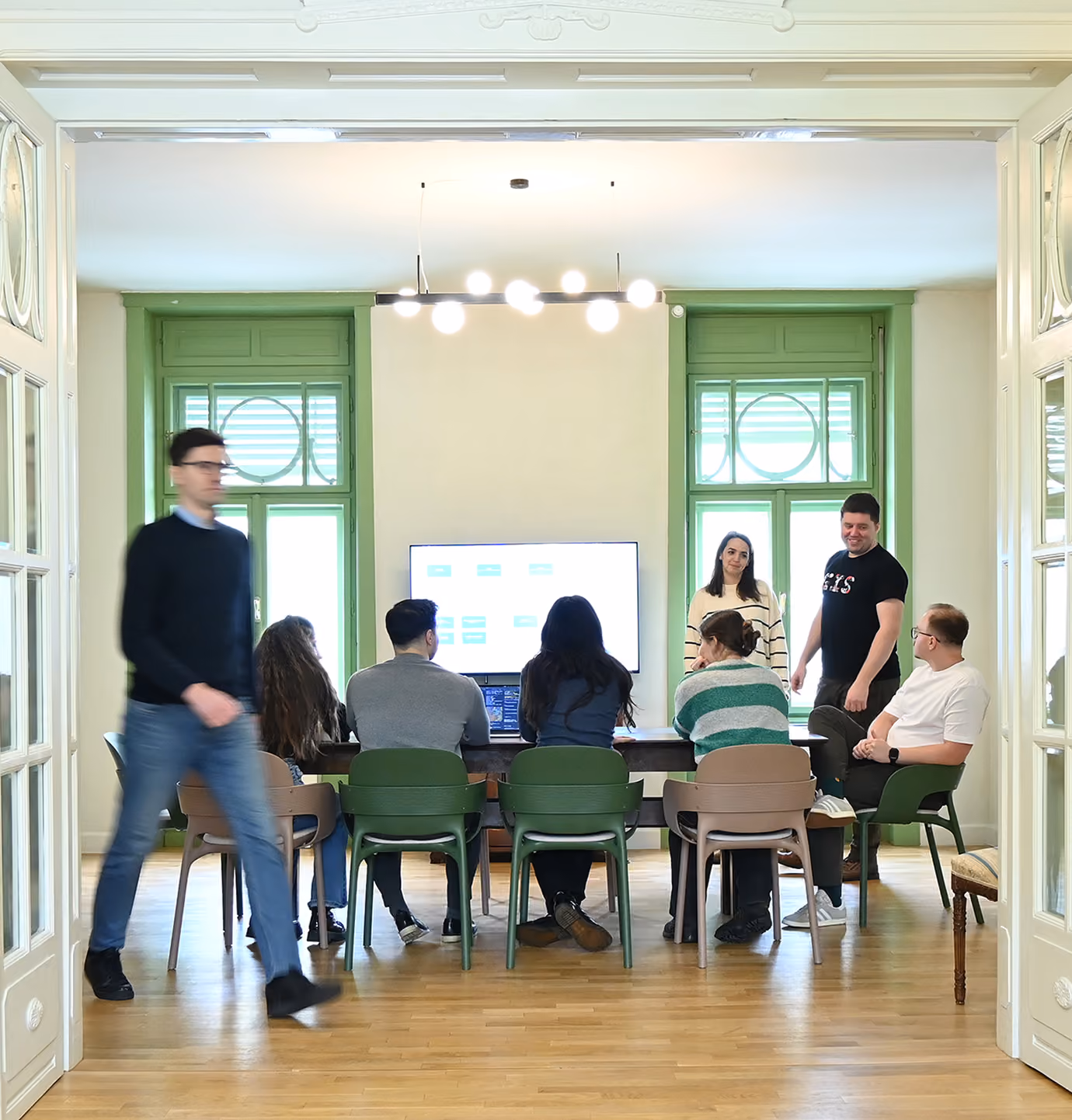 A group of professionals in a brightly lit meeting room with large windows framed in green, gathered around a big table as they discuss ideas.