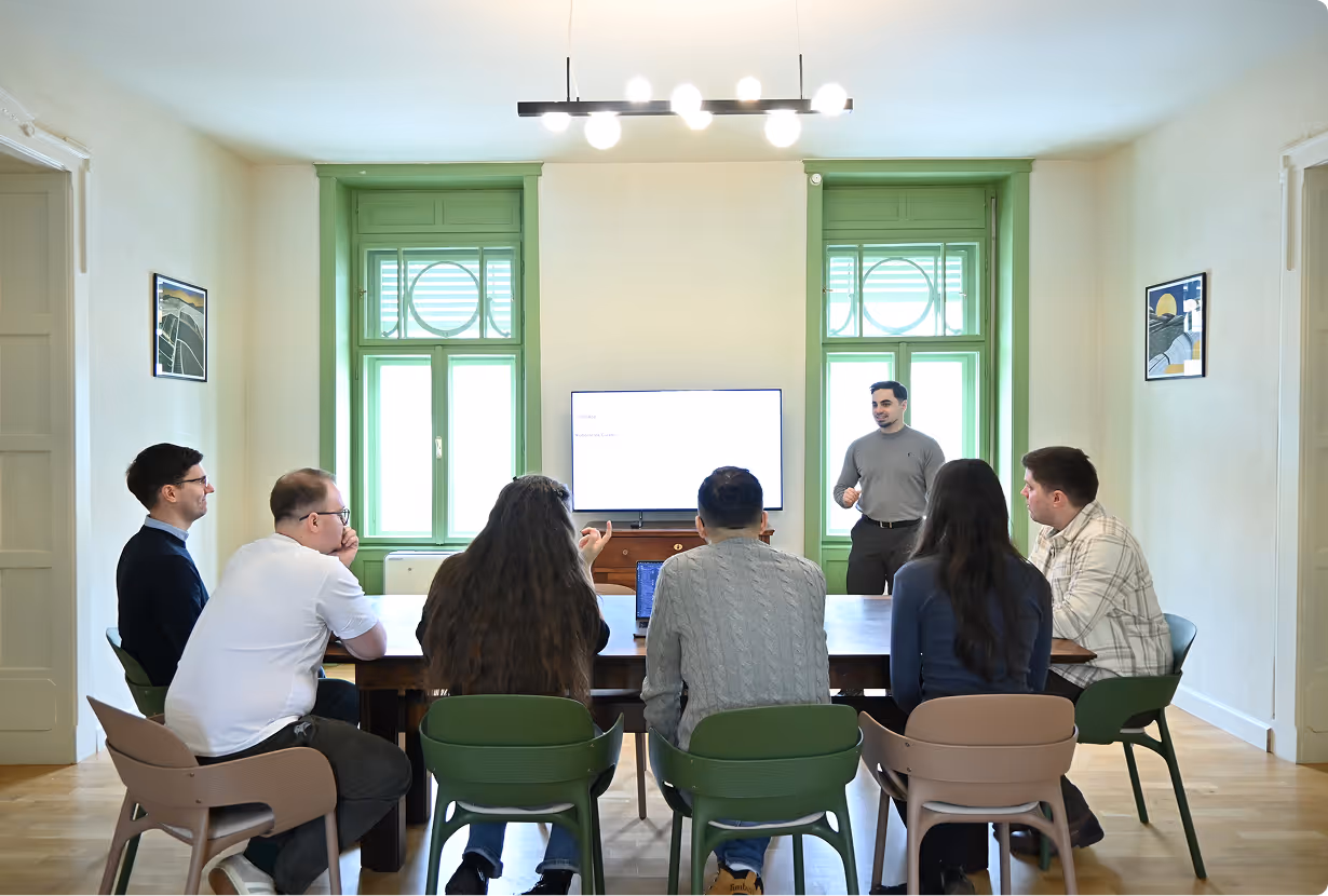 A group of people sitting around a conference table in a meeting room with large green windows, watching a presenter and a bright screen.