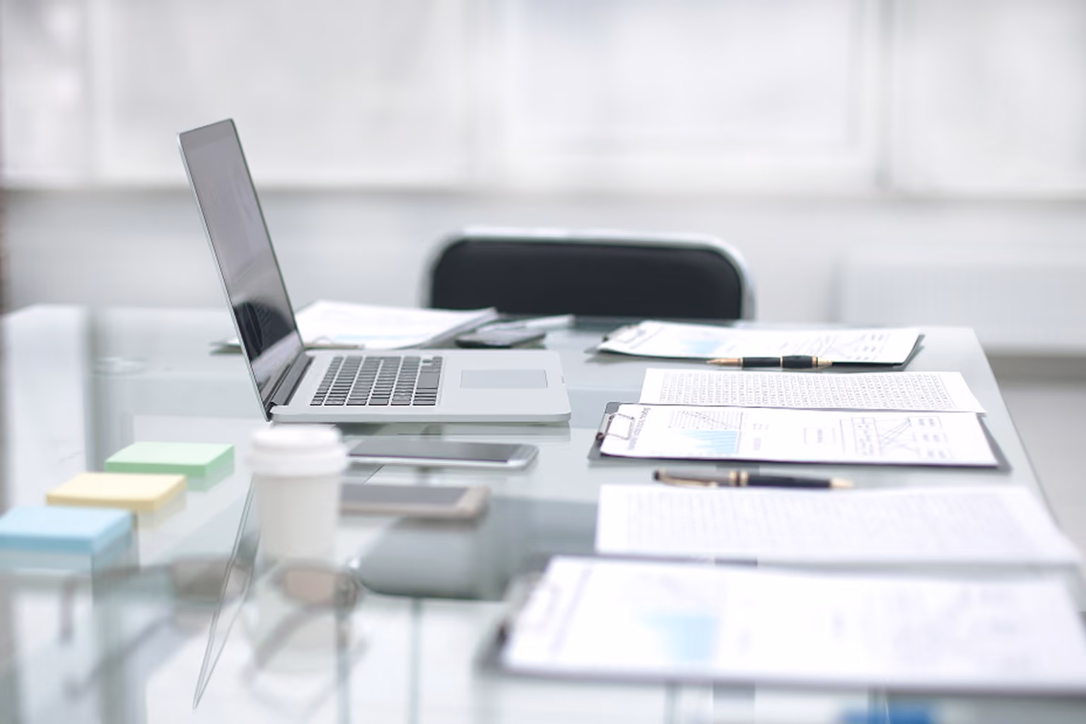 Glass office desk with an open laptop, multiple clipboards holding documents, pens, smartphone, colorful sticky notes, and a coffee cup.