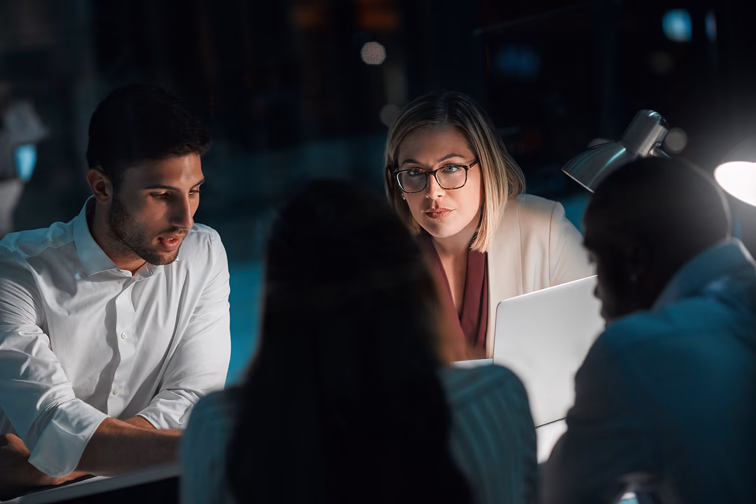 Four diverse colleagues gathered around a laptop at a desk in a dimly lit office, engaged in a focused discussion.