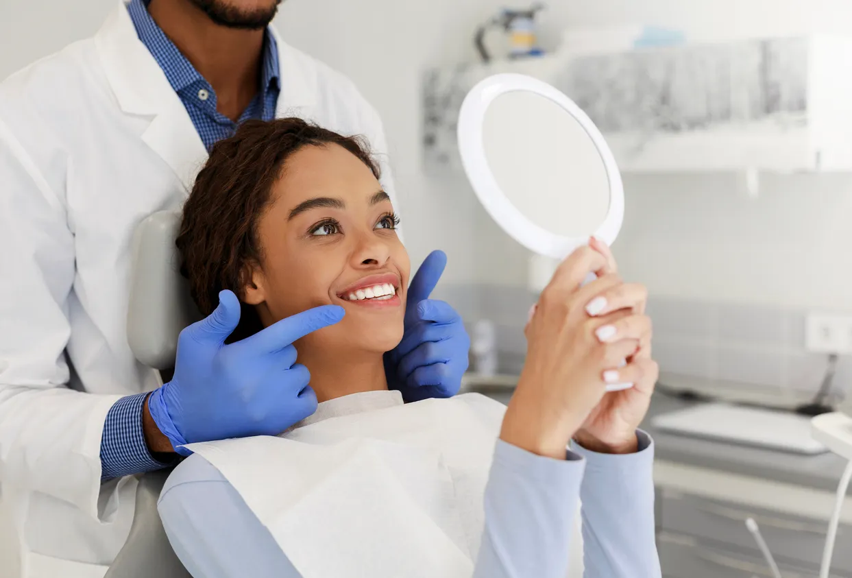 A woman smiles while holding a mirror, with a dentist standing beside her wearing gloves.