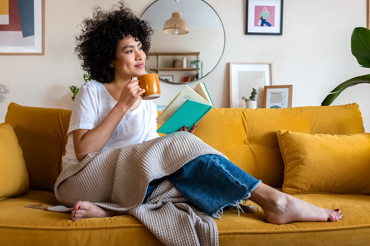 Person sitting on a yellow couch, drinking from a mug, and reading a book.
