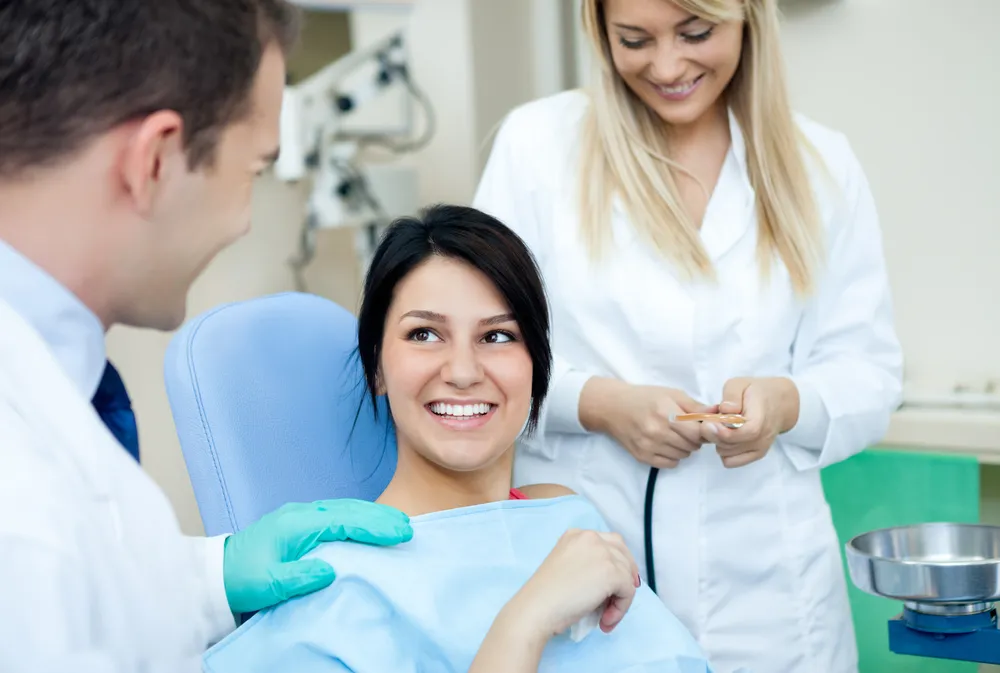 A woman sits in a dental chair, smiling at a dentist, with an assistant nearby.