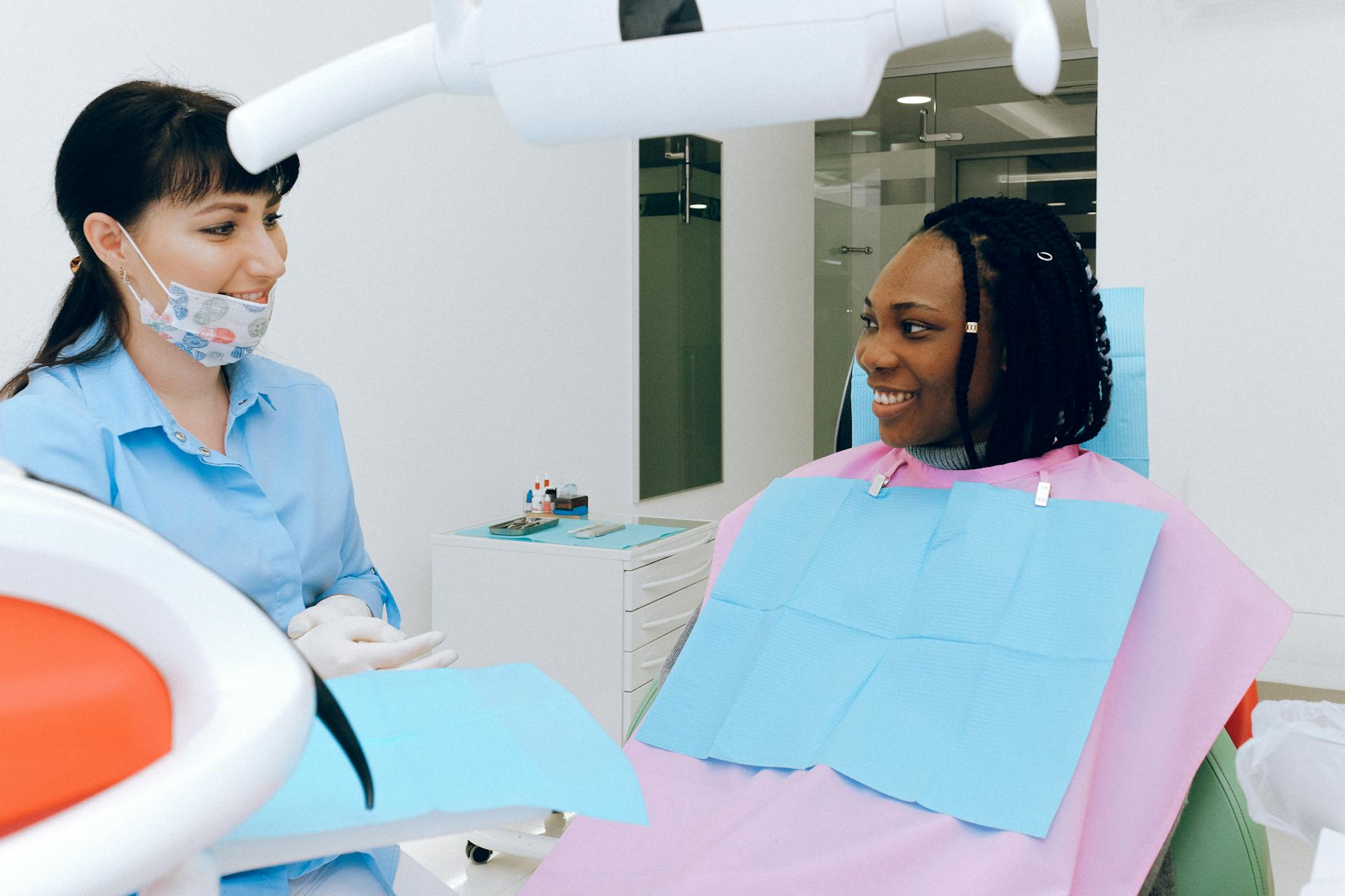 A dentist talks to a patient sitting in a dental chair, both are smiling.